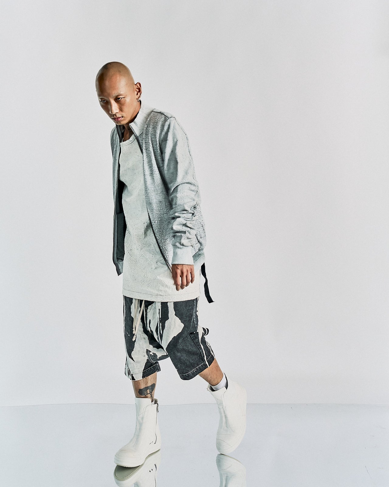 Person in a minimalist, edgy Rick Owens outfit featuring a light gray textured jacket, long white shirt, abstract print shorts, and white boots. Calm expression.