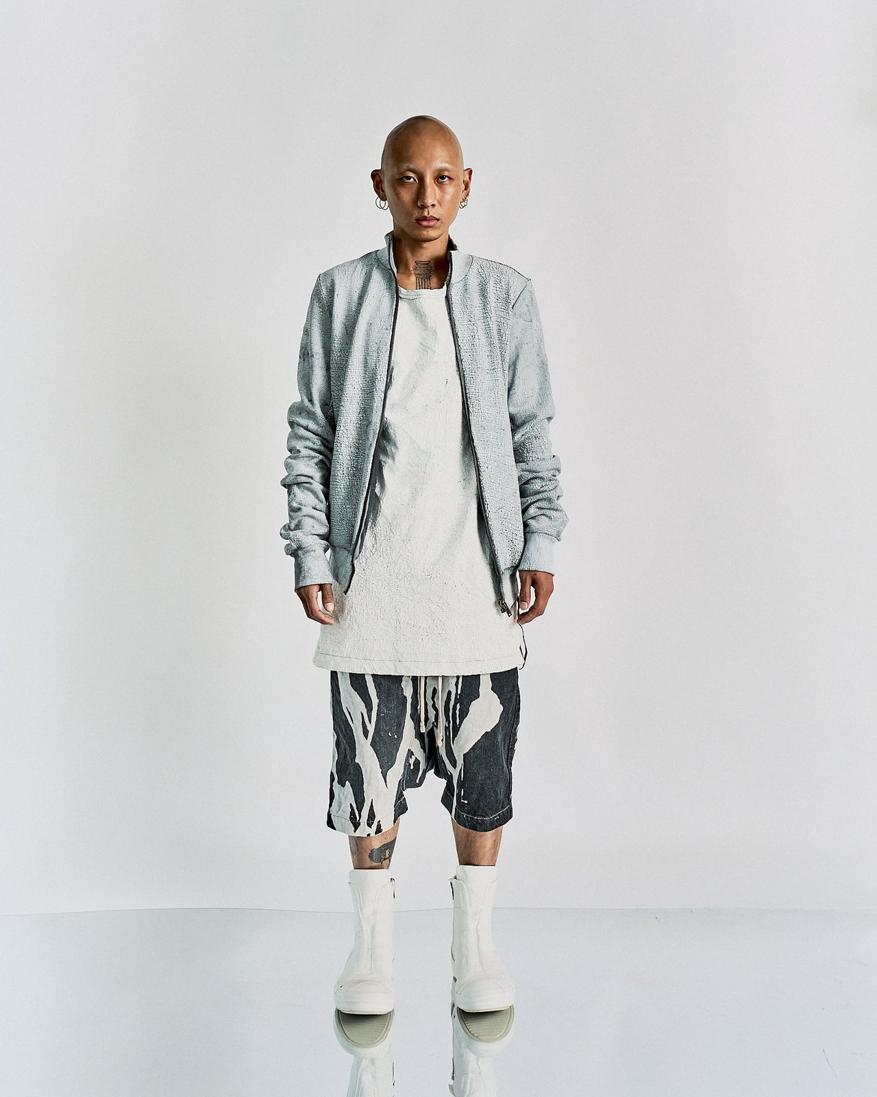 Person in a minimalist, edgy Rick Owens outfit featuring a light gray textured jacket, long white shirt, abstract print shorts, and white boots. Calm expression.