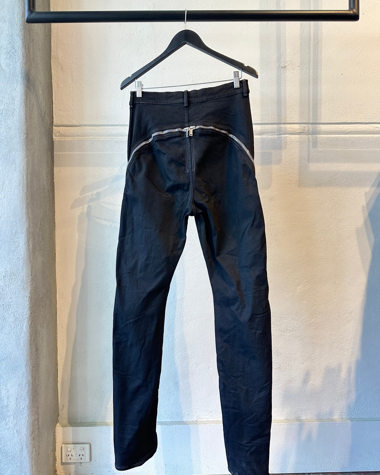 Black Rick Owens jeans hanging on a hanger against a light-colored wall.