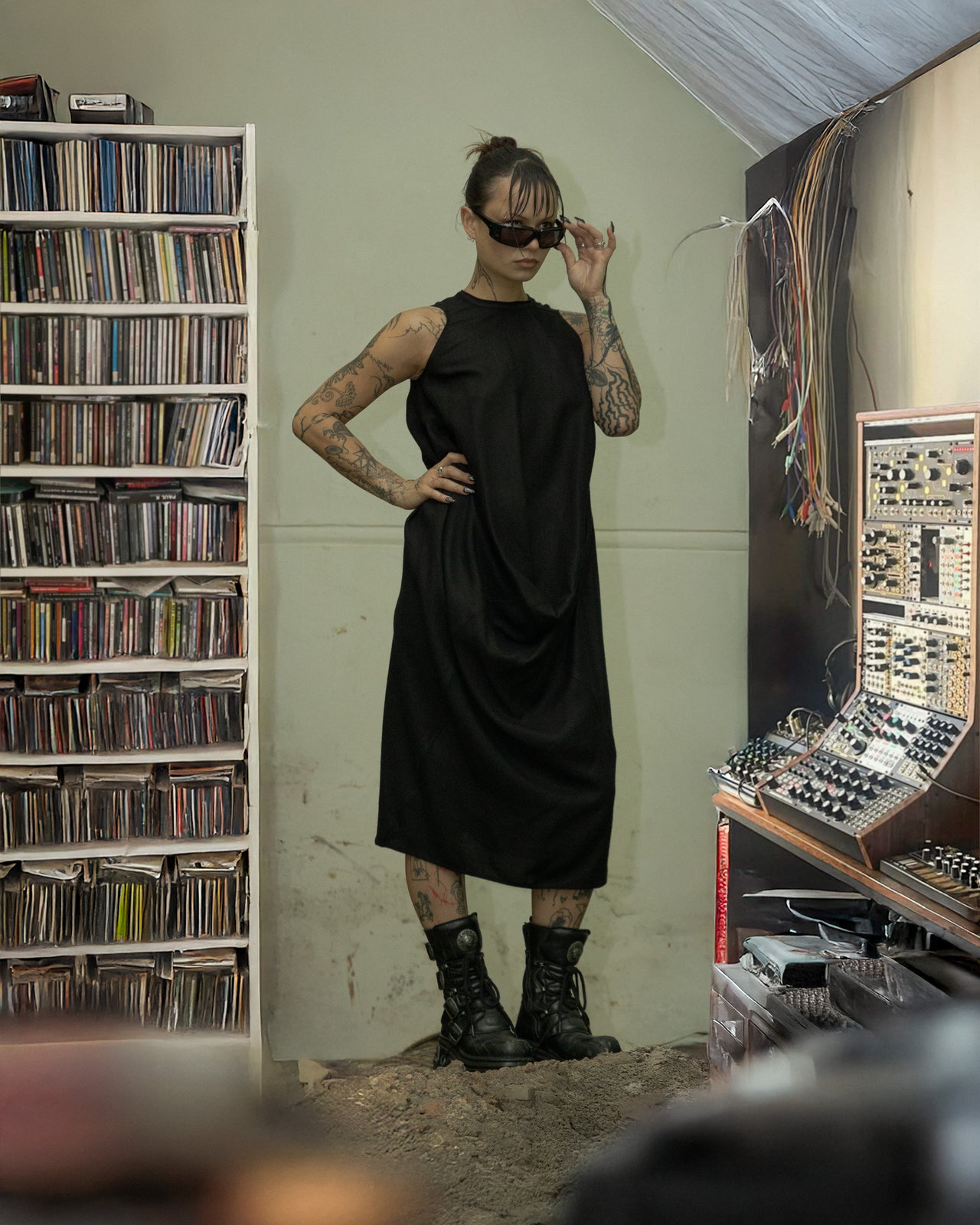 A person standing in a room with audio equipment and bookshelves, wearing a black sleeveless Rick Owens A-line dress with a protruding front detail and black boots.