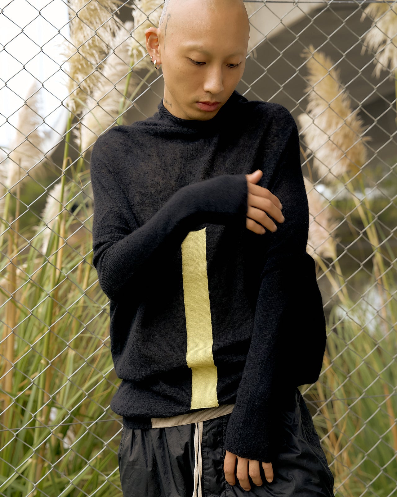 A person wearing a black long-sleeve Rick Owens sweater with geometric intarsia cotton patches and a ribbed collar, paired with black shorts and sneakers, standing near a wire fence with plants in the background.