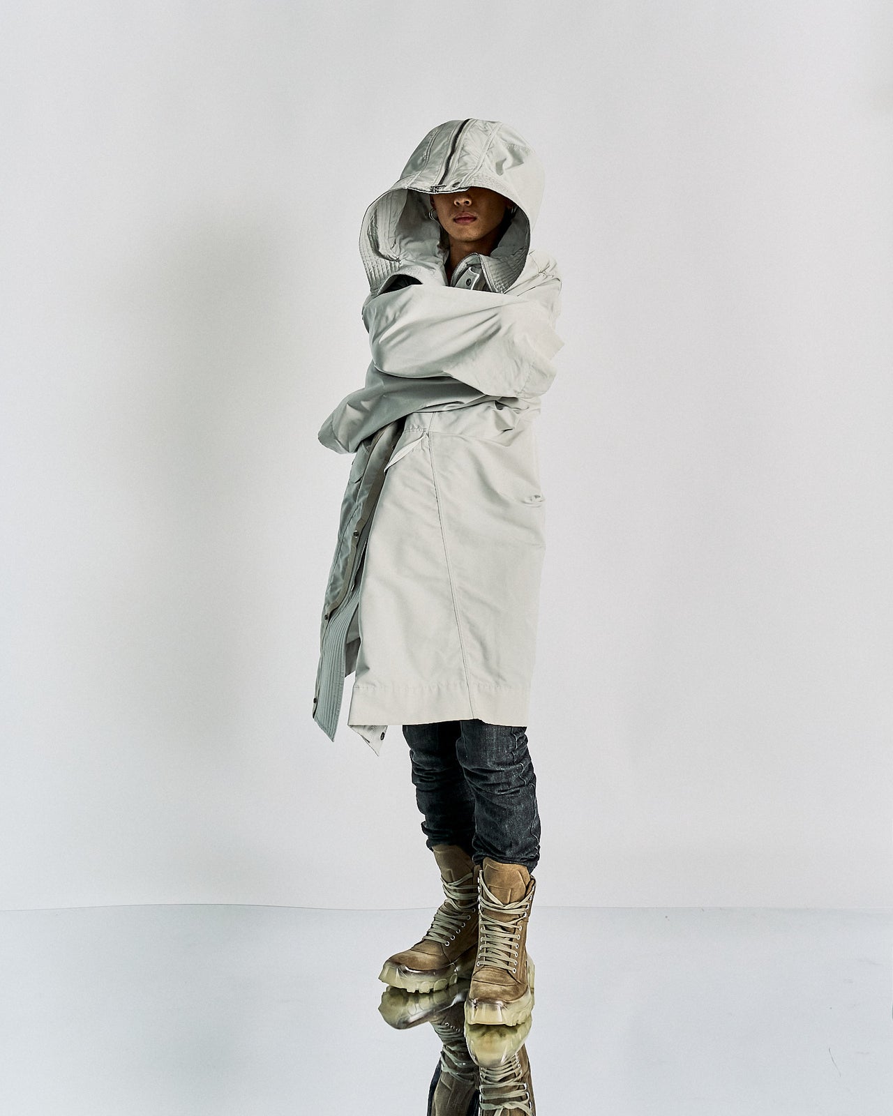 A person wearing an oversized grey hooded Rick Owens parka with a zipper and a pair of grey boots with yellow accents. The photo is monochrome.