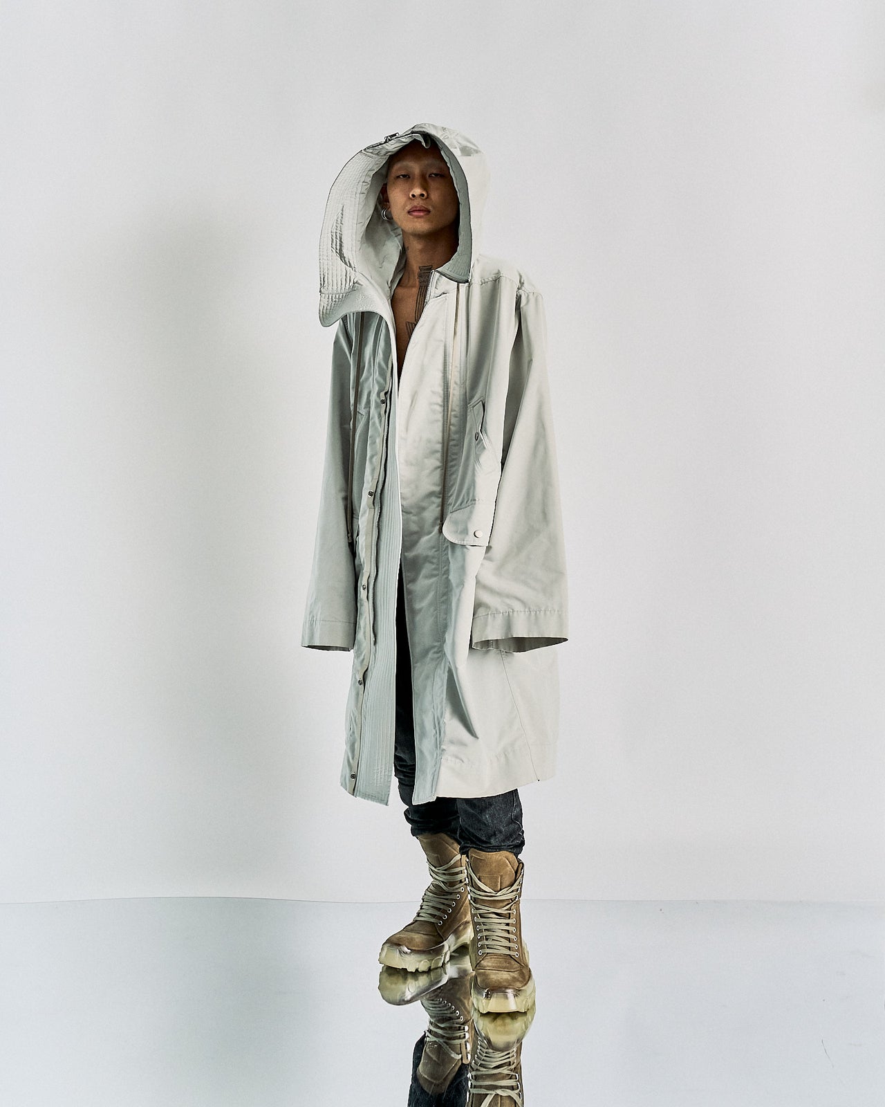 A person wearing an oversized grey hooded Rick Owens parka with a zipper and a pair of grey boots with yellow accents. The photo is monochrome.