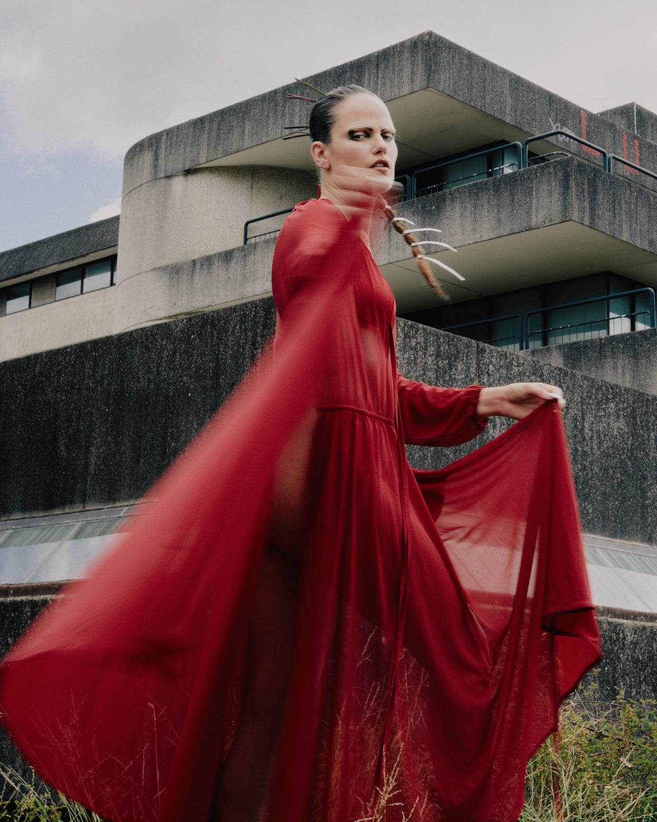 Model walks confidently on runway in vibrant sheer red Vetements dress and black boots, holding a blue clutch. Audience watches attentively in background.