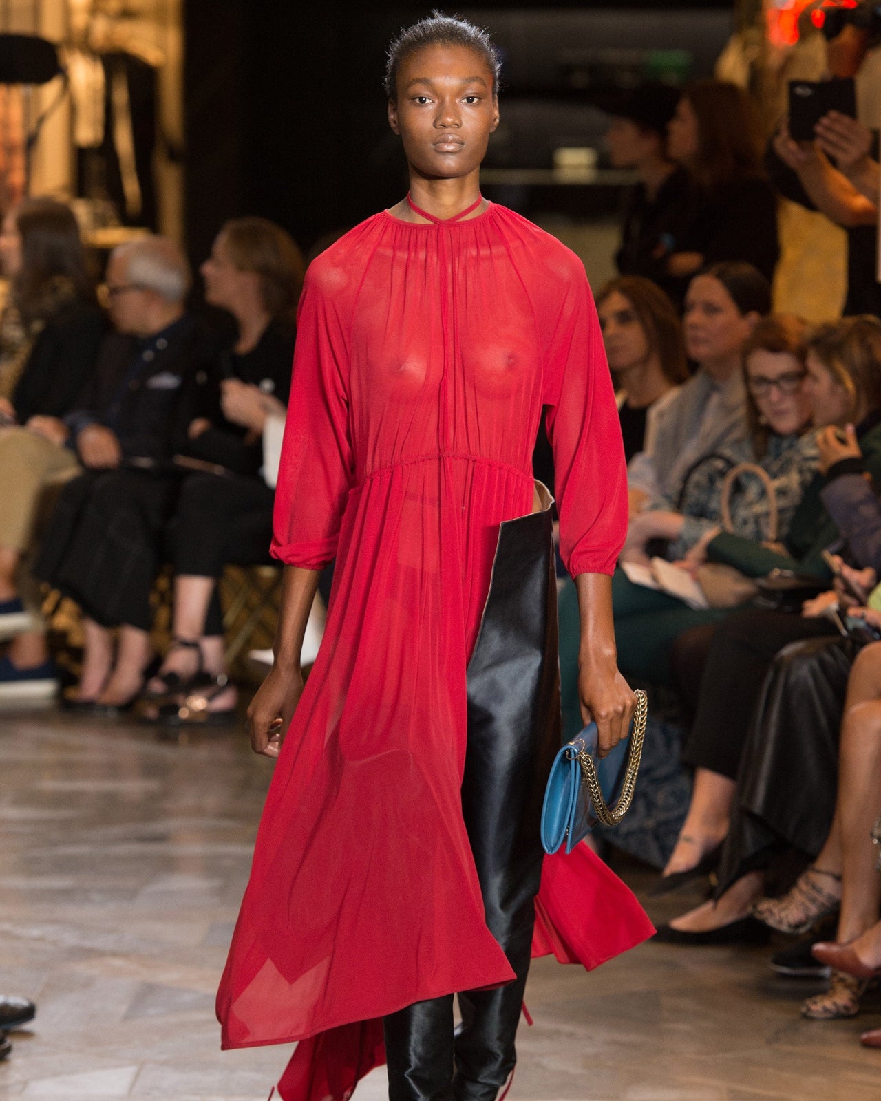 Model walks confidently on runway in vibrant sheer red Vetements dress and black boots, holding a blue clutch. Audience watches attentively in background.
