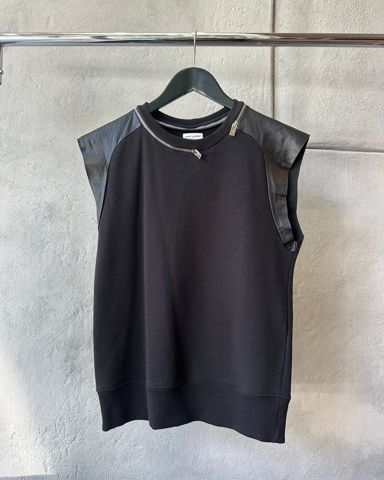 Black sleeveless Saint Laurent top with leather sleeves on a hanger against a gray wall.