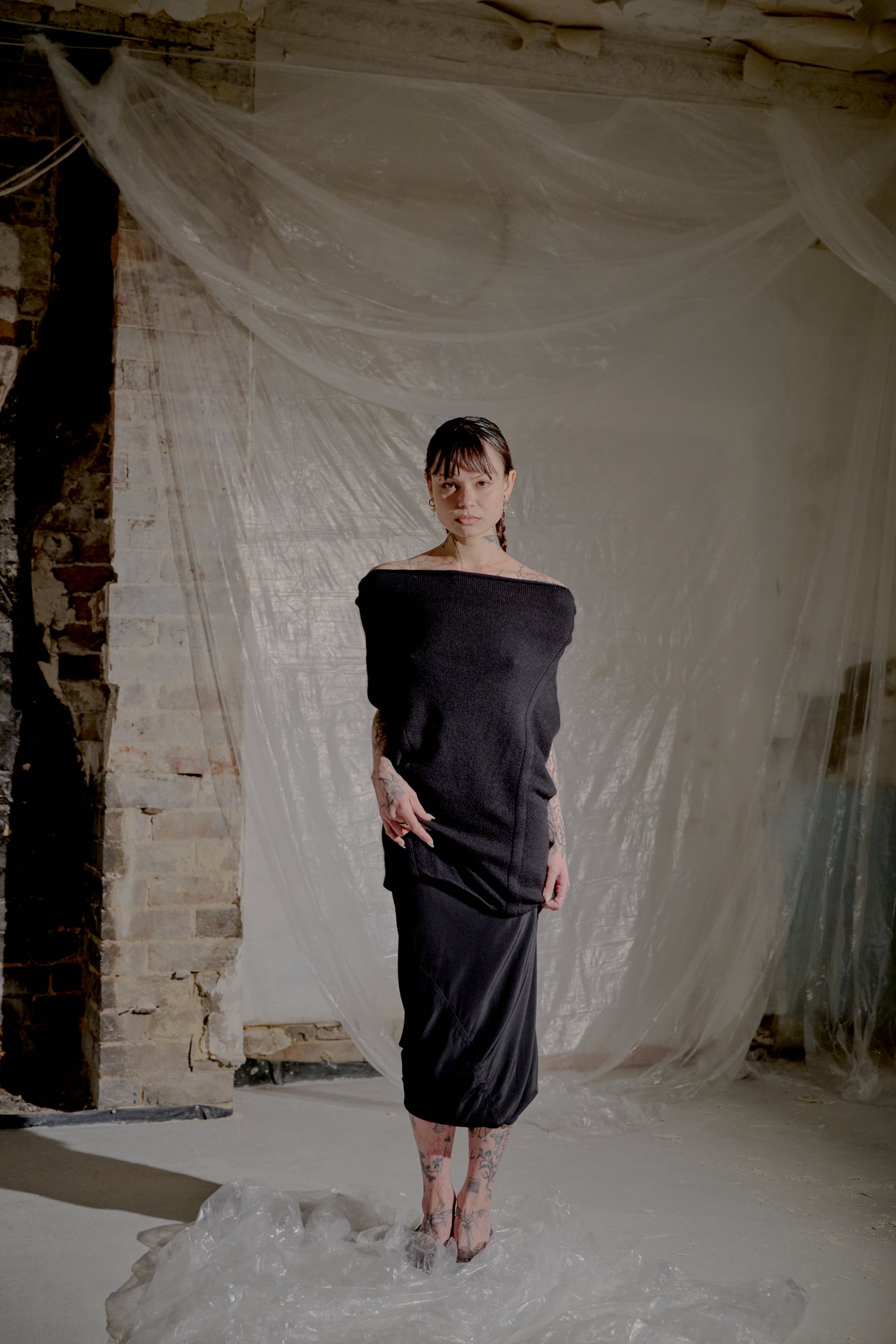 Woman in a black off-shoulder Saint Laurent dress standing in a room with white fabric draped on the wall.