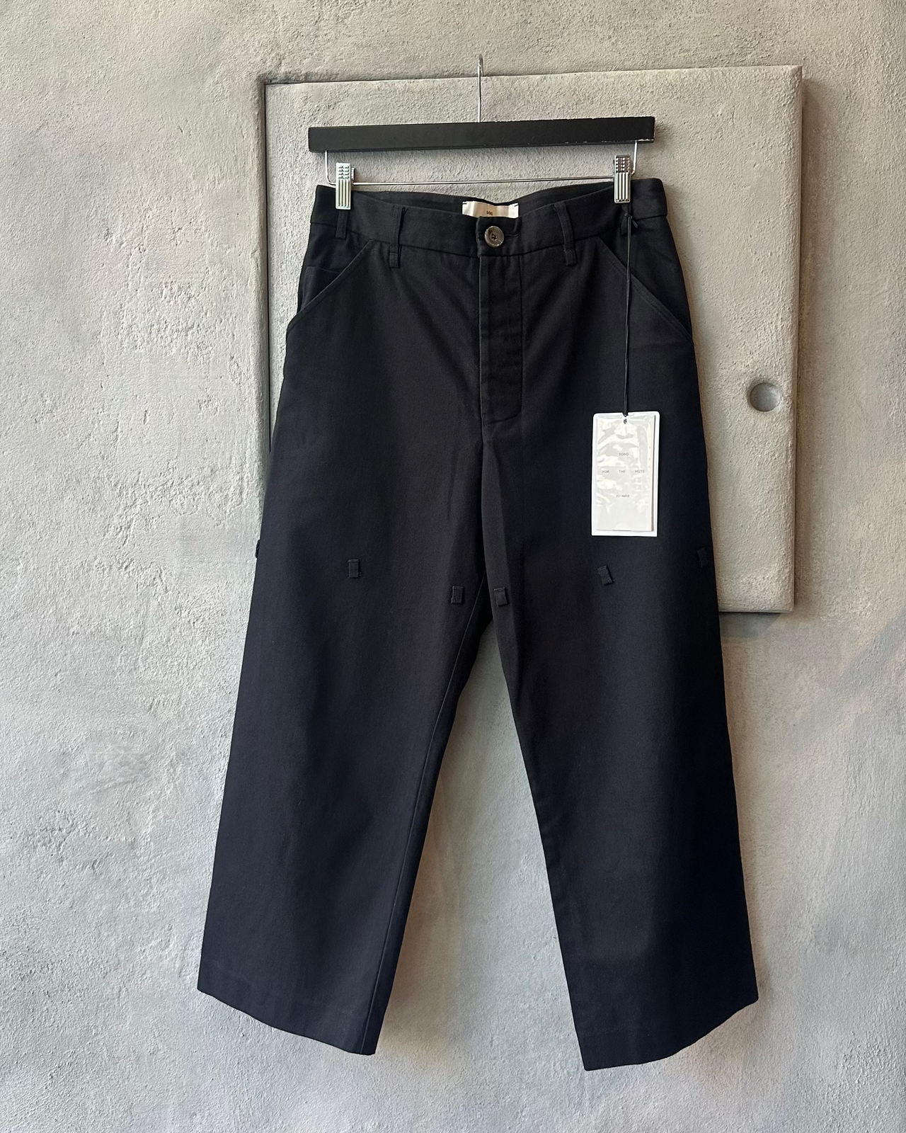 Black Song For The Mute pants hanging on a rack against a gray concrete wall