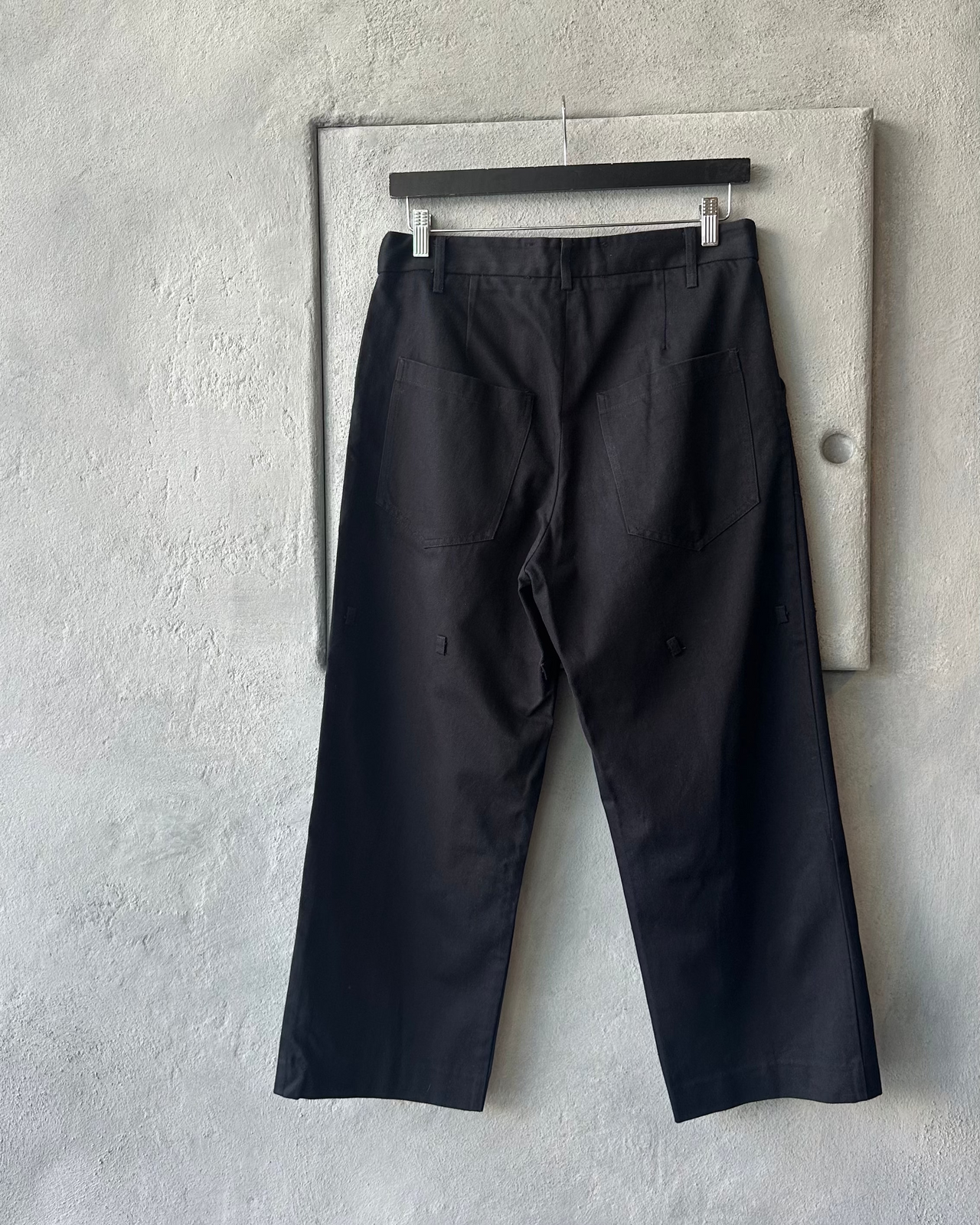 Black Song For The Mute pants hanging on a rack against a gray concrete wall