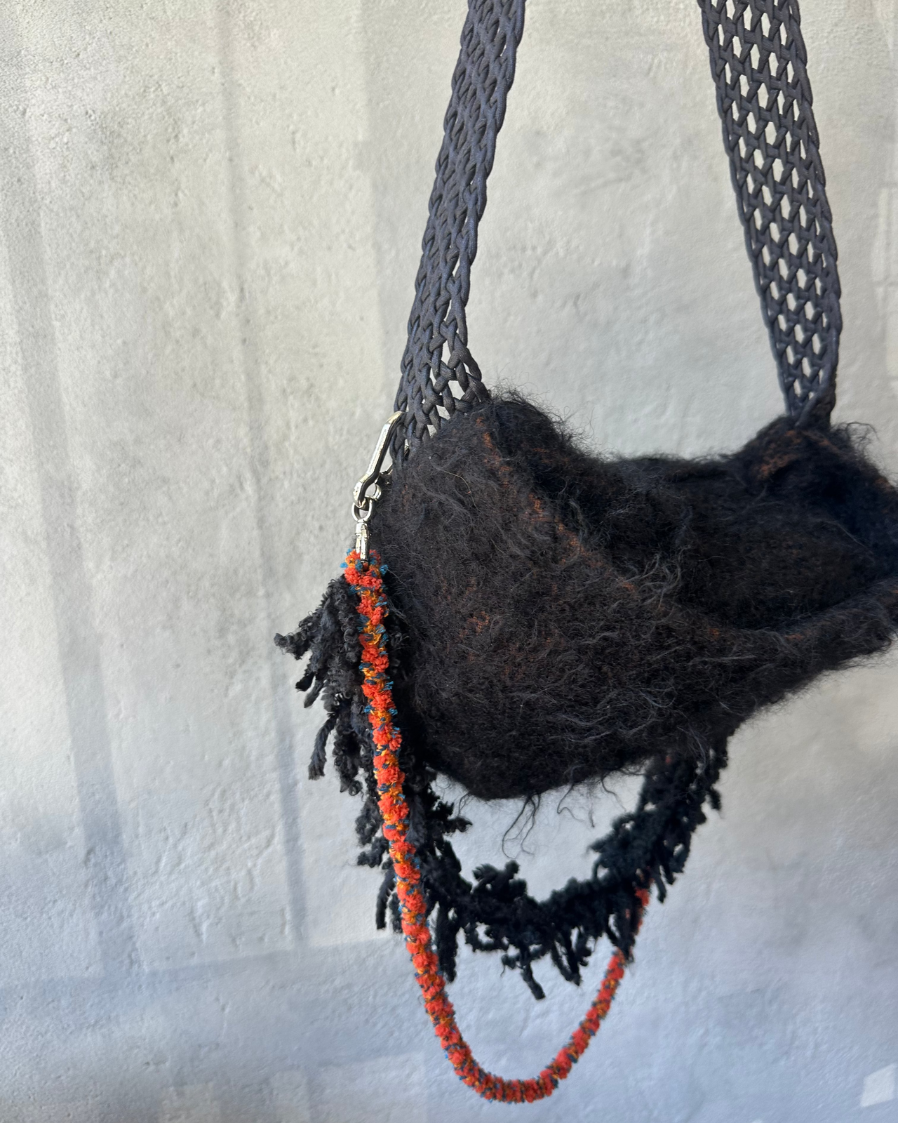 Black woven Song For The Mute bag with red rope hanging against a textured wall.
