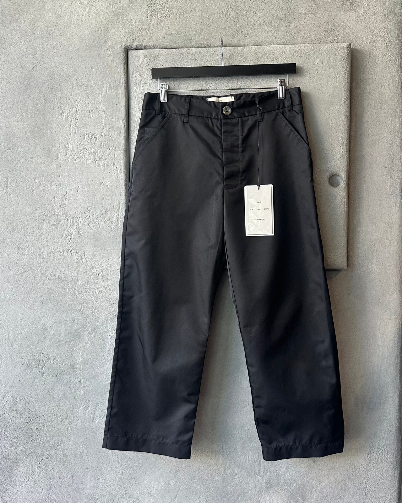 Black Song For The Mute pants with a tag on a gray concrete wall