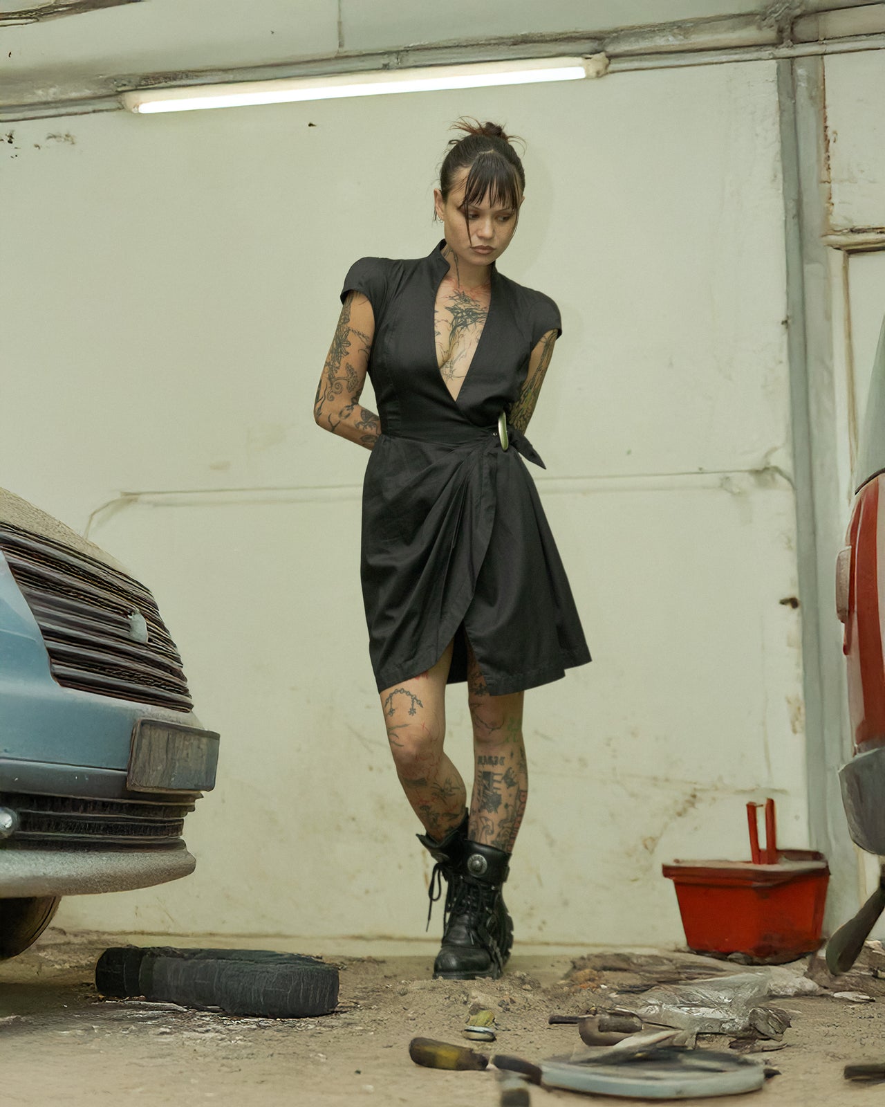 A person standing in a garage wearing a black belted Thierry Mugler dress with cap sleeves, paired with black boots.
