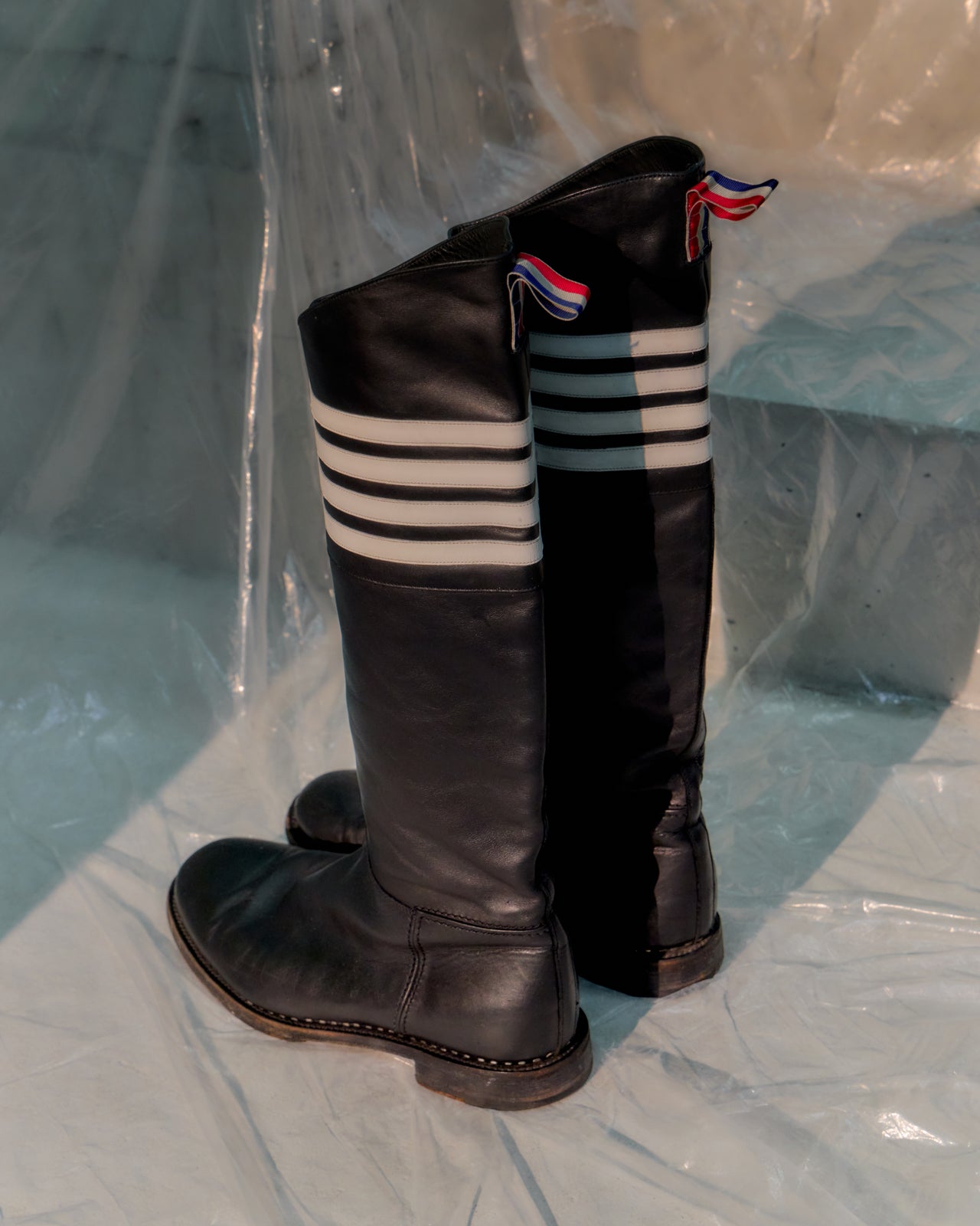 Black knee-high Thom Browne boot with three white stripes on a light blue background