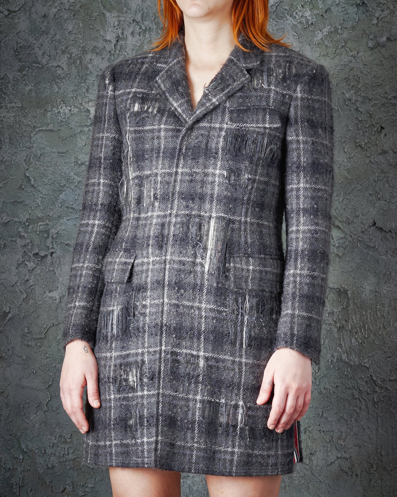 A person with straight red hair stands indoors against a textured gray wall, wearing the FW 2016 Distressed Tartan Mohair Coat by Thom Browne as a grey plaid blazer dress paired with black chunky boots.
