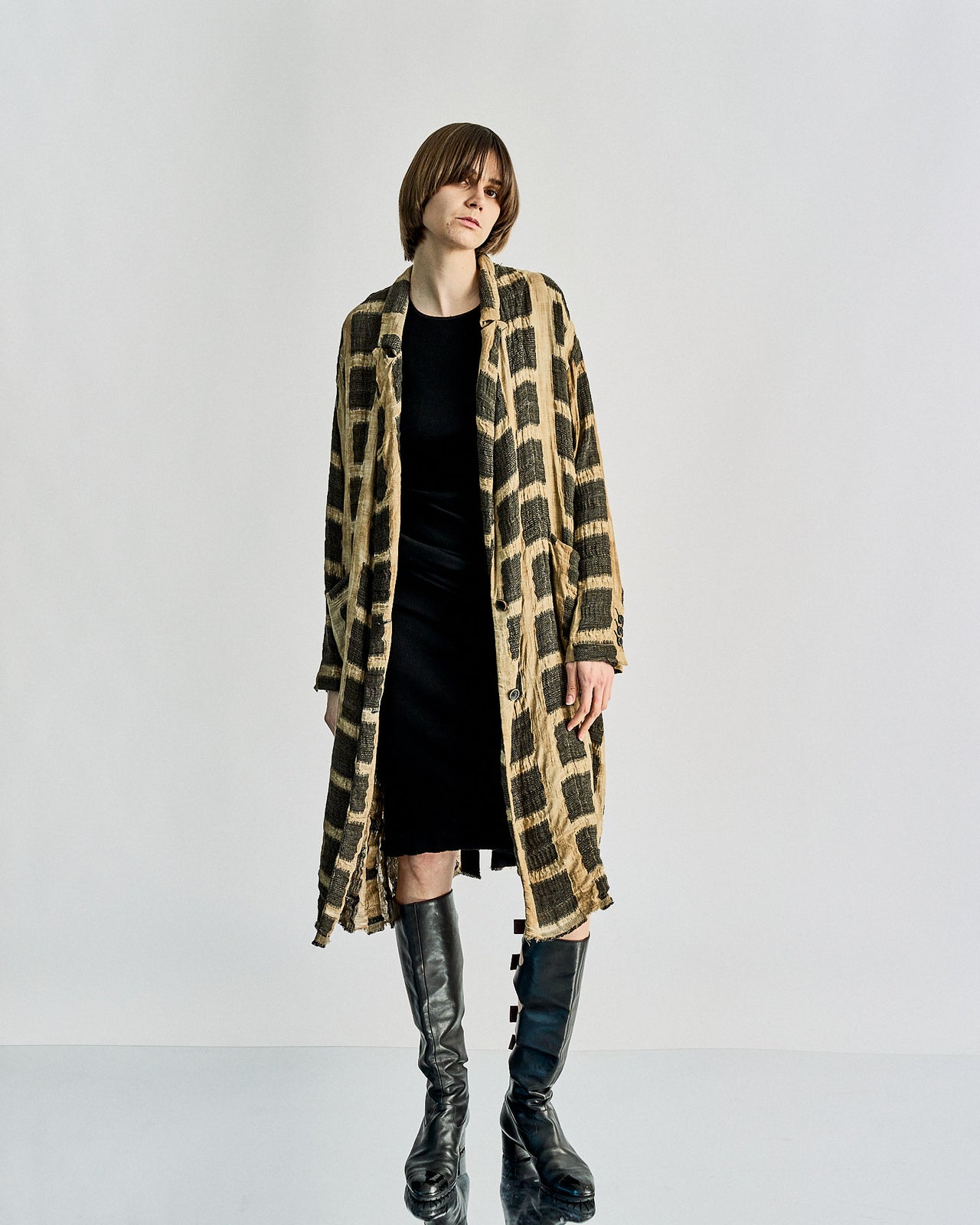 A longline Uma Wang coat in a textural linen with a black and white animal pattern, featuring a slim sculptural collar, two-button front, and pockets at the hips. The coat is on a mannequin and the model is wearing a black dress and black boots.