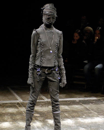 Person wearing a dark, textured Undercover outfit with gloves and boots on a dark background