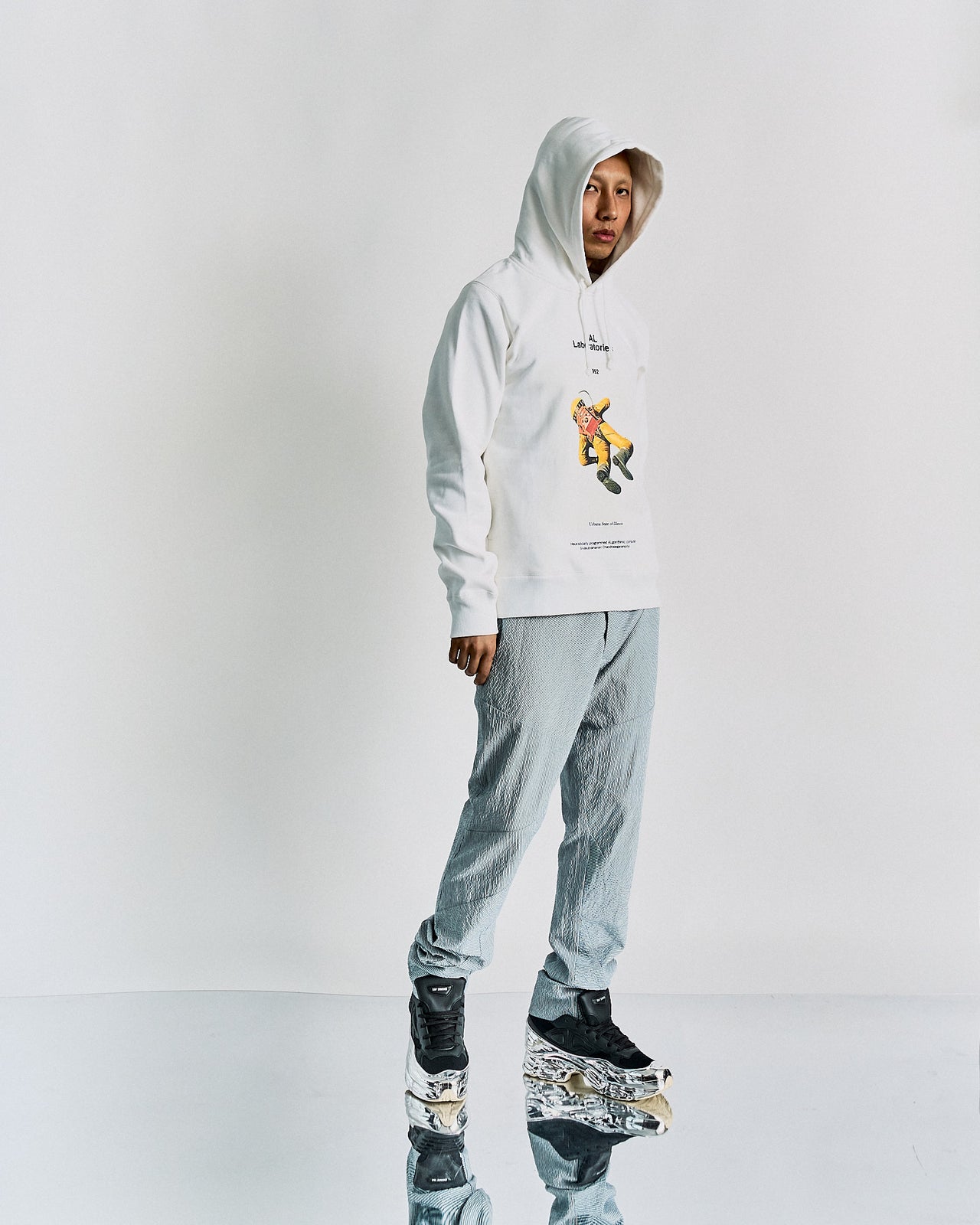 A person standing in a studio setting, wearing a white Undercover hoodie with a vintage-inspired 2001: A Space Odyssey print and gray pants.