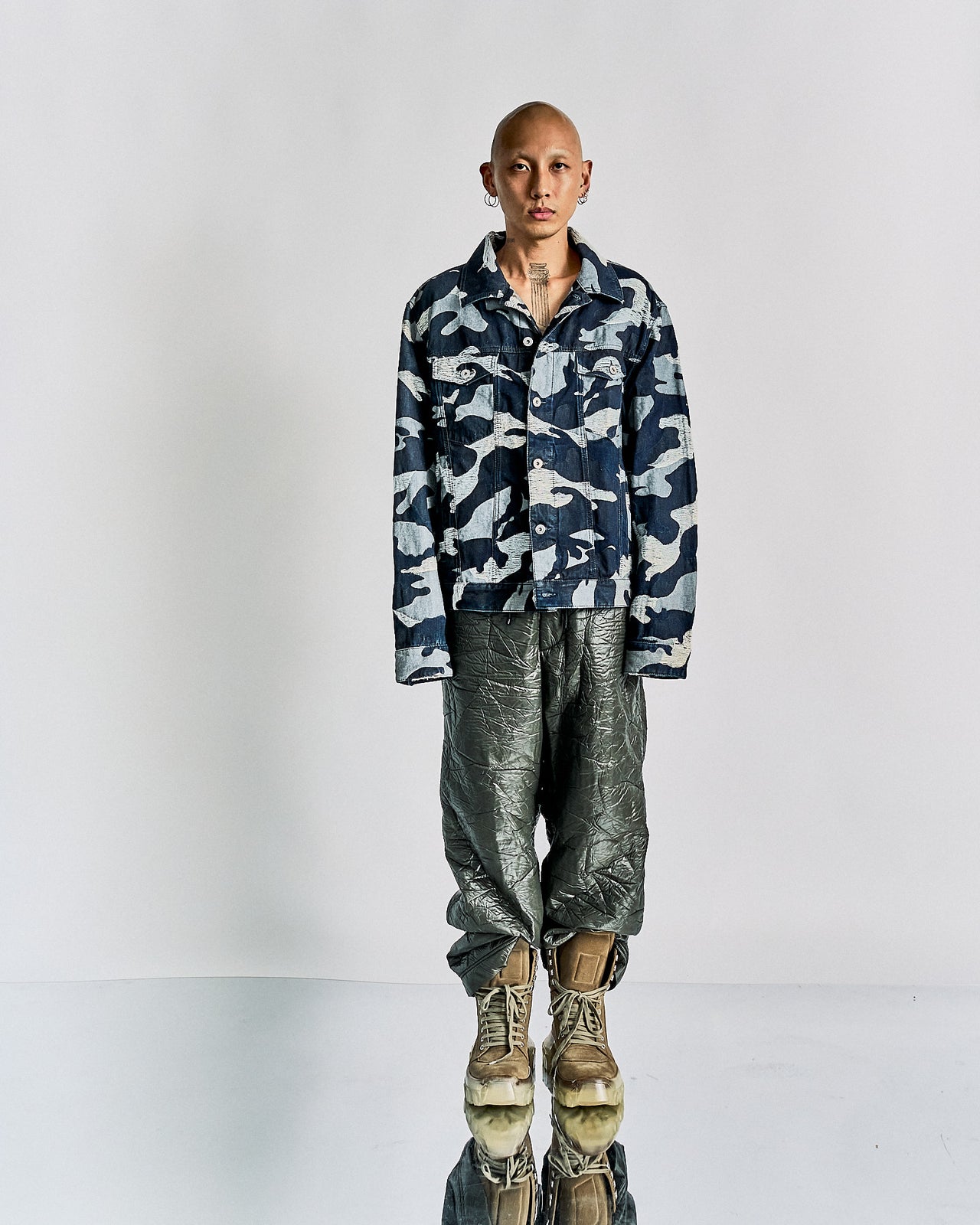 A person with a shaved head wears a blue camouflage Valentino jacket, metallic textured pants, and tan boots, standing confidently against a plain backdrop.