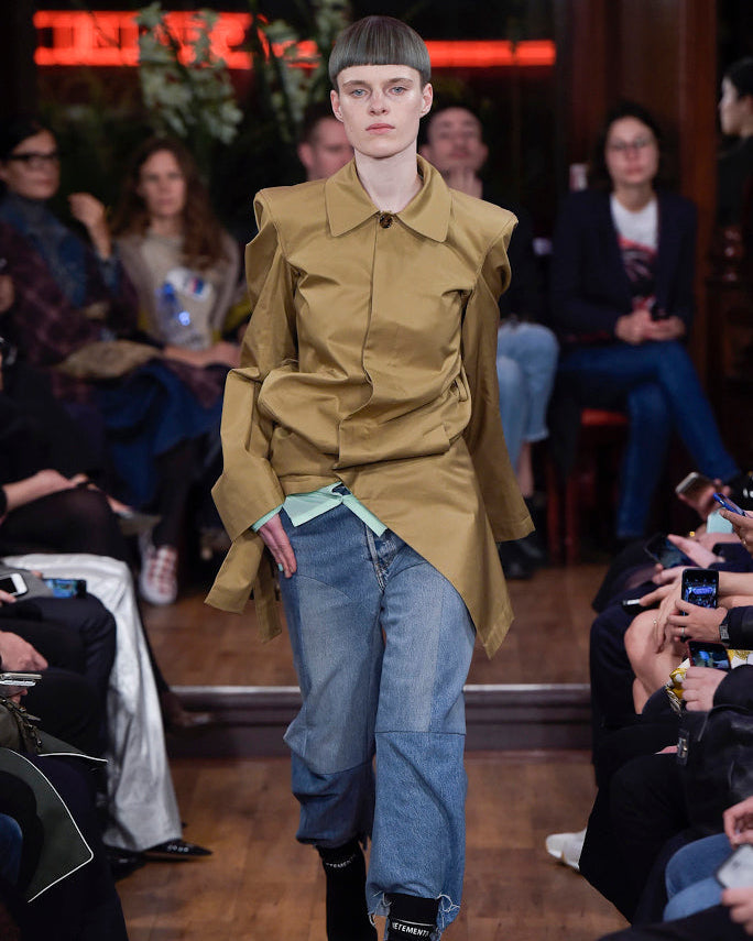 A model walks the runway in the SS 2016 Structured trench coat from Vetements with angular shoulders, layered over a mint top and baggy blue jeans, channeling Demna Gvasalia's Spring/Summer 2016 style as the audience snaps photos.