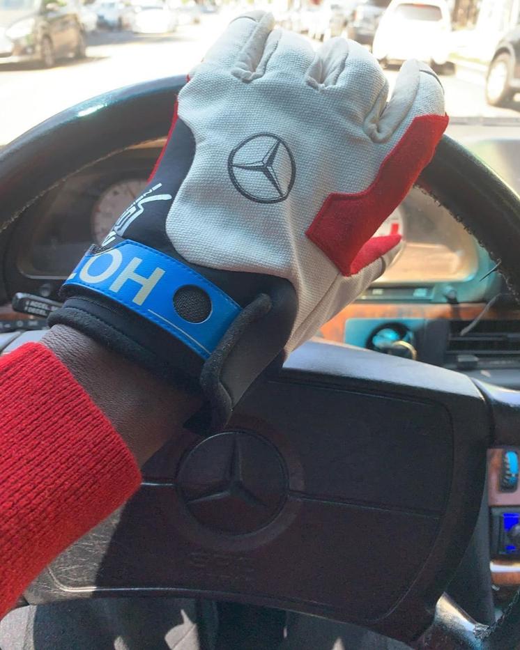 A person's hand wearing a white and red driving glove from Virgil Abloh with the Mercedes Benz logo on the back, resting on a car's steering wheel.