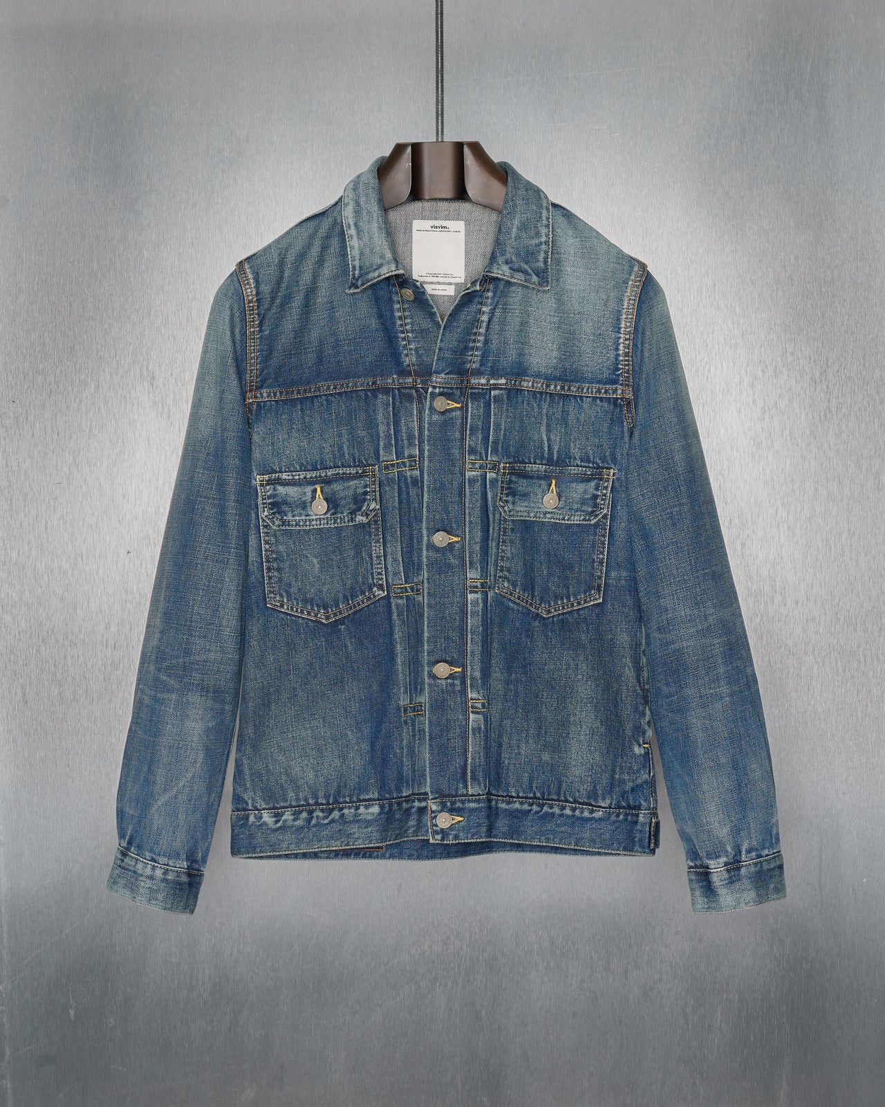 A blue denim Visvim jacket with a faded, worn-in look is hanging on a black hanger against a metallic textured background. The jacket has two chest pockets.