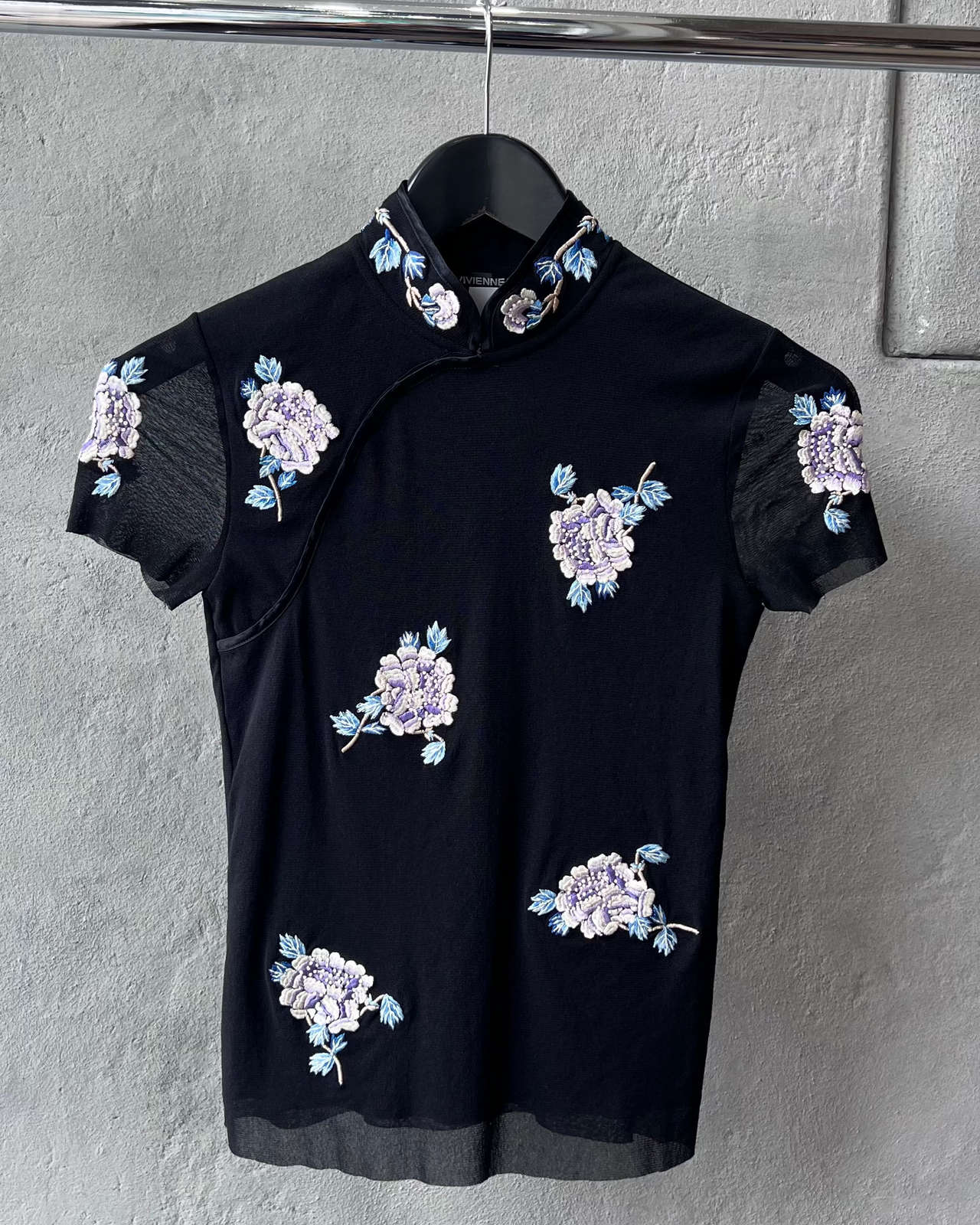 Black Vivienne Tam blouse with floral embroidery on a hanger against a gray wall.