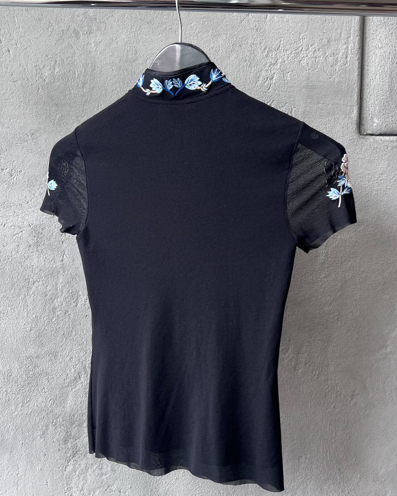 Black Vivienne Tam blouse with floral embroidery on a hanger against a gray wall.