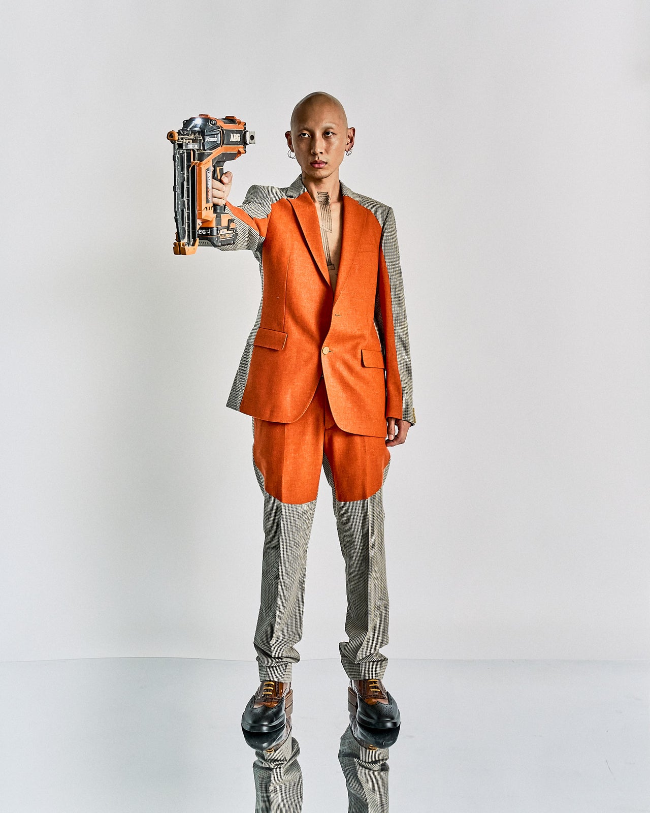 A model on a runway wears a Walter Van Beirendonck outfit with an orange blazer and shorts over checkered pants, accessorised with a matching orange bowtie, mask, and hat.