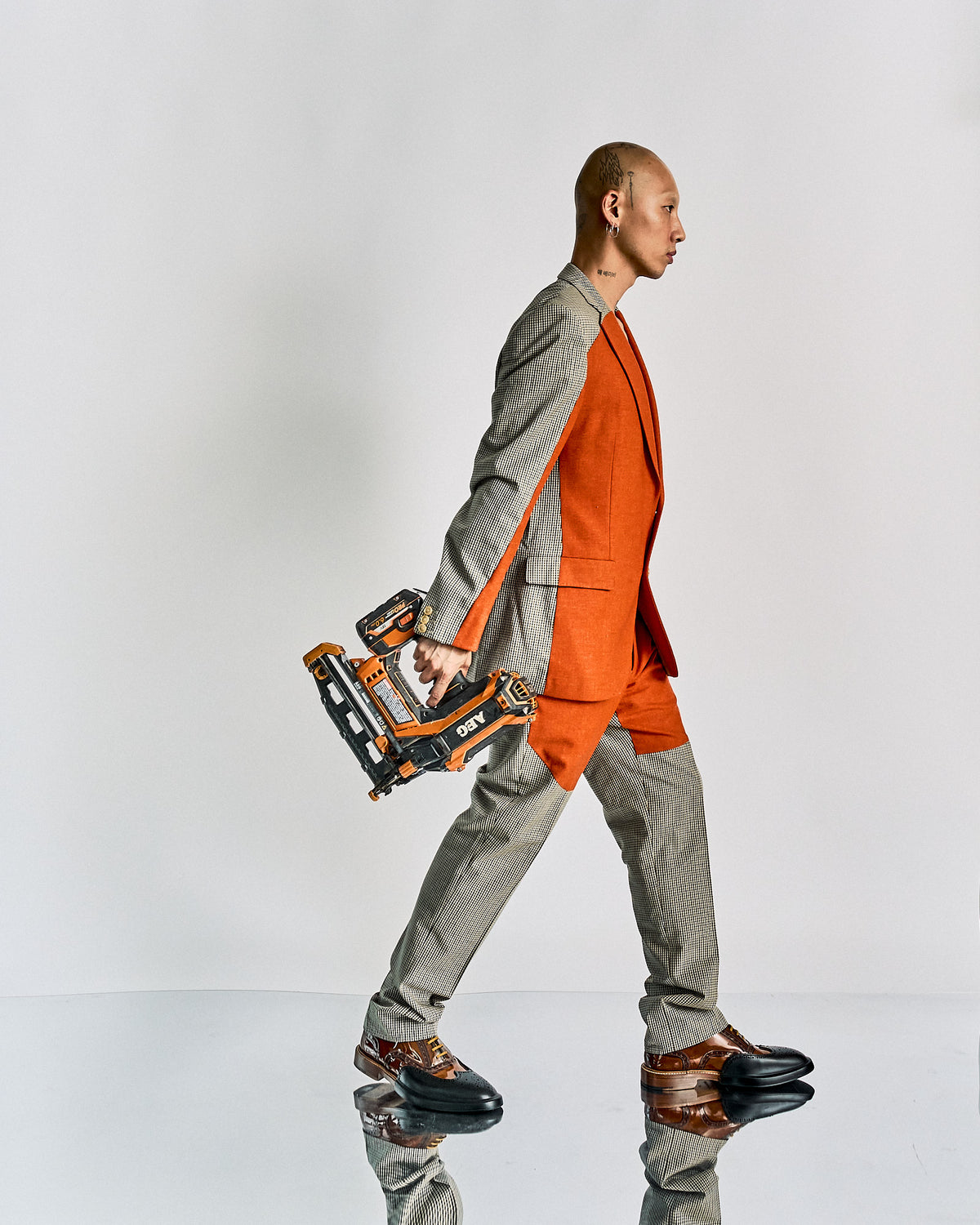 A model on a runway wears a Walter Van Beirendonck outfit with an orange blazer and shorts over checkered pants, accessorised with a matching orange bowtie, mask, and hat.