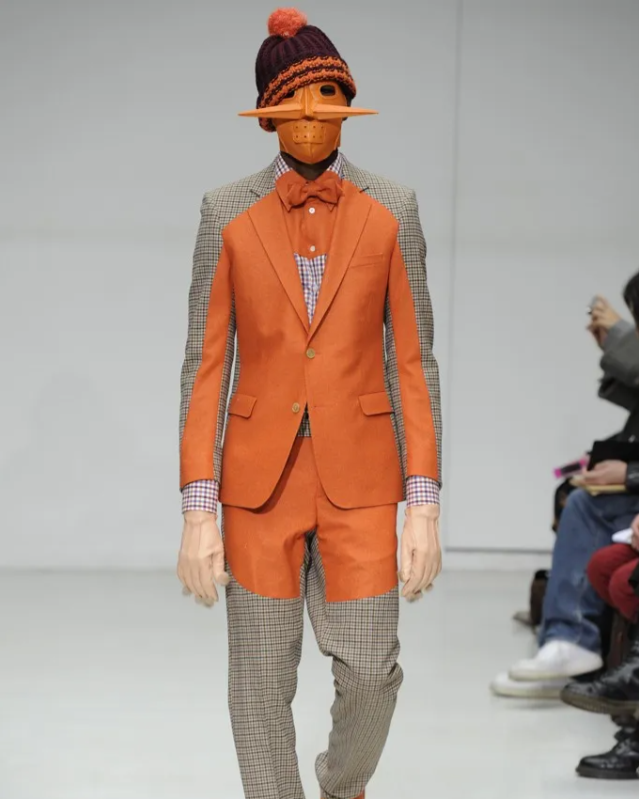 A model on a runway wears a Walter Van Beirendonck outfit with an orange blazer and shorts over checkered pants, accessorised with a matching orange bowtie, mask, and hat.