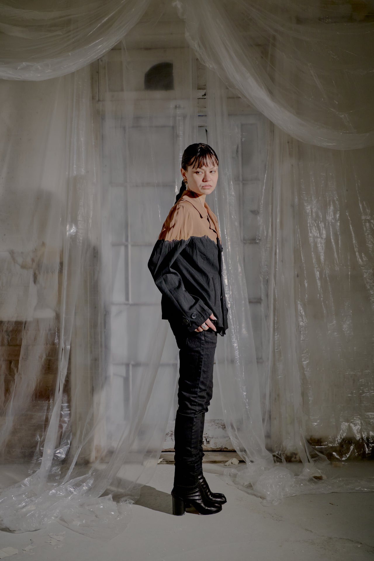 Person wearing a black and brown Y's by Yohji Yamamoto jacket over a beige shirt, standing in a room with draped fabric.
