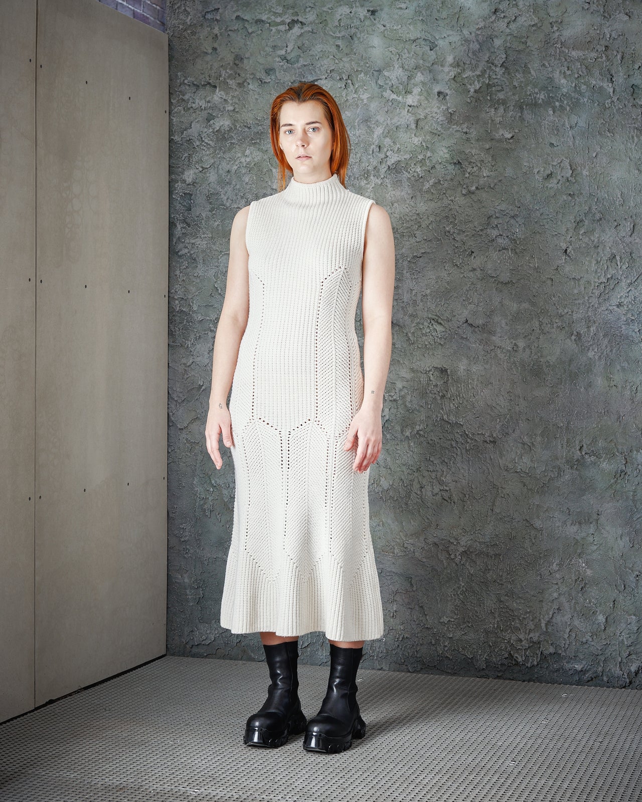 A model wearing a sleeveless white cable knit Alexander McQueen midi dress with a high neck and a soft fish tail skirt with rolled hem, paired with black boots.