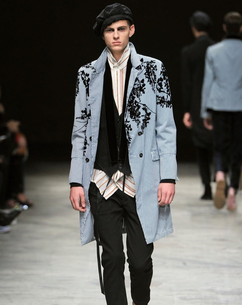 A male model walks down the runway in a light blue Ann Demeulemeester trench coat with black floral patterns, layered over a striped shirt and black pants. He wears a black beret.