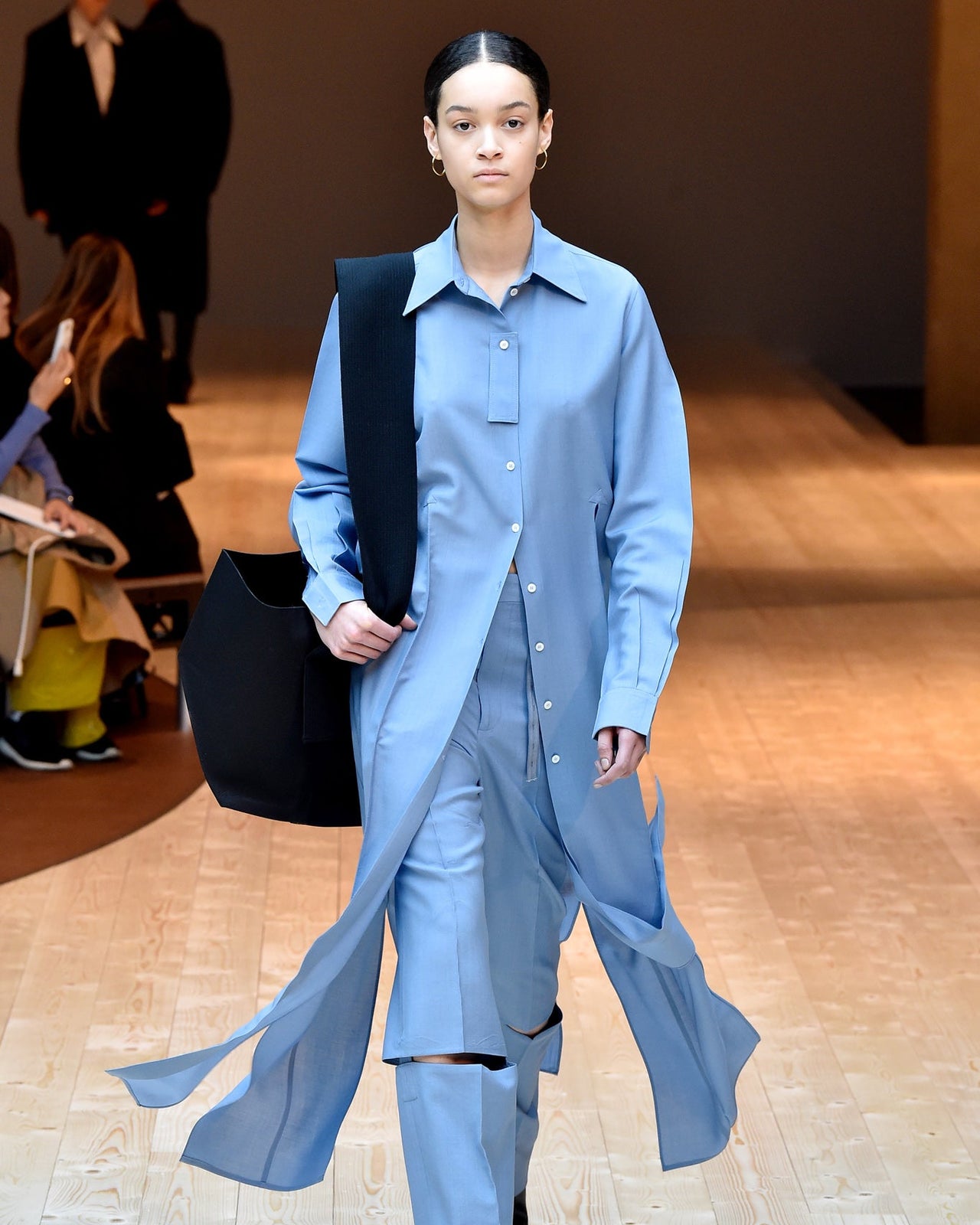 Model on a runway wears a flowing, light blue Celine tunic and pants, holding a black tote. The setting is bright and minimalist, with a neutral-toned background.