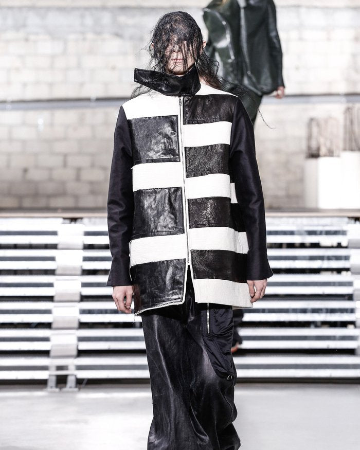 Model in avant-garde fashion walks runway wearing oversized black and white striped Rick Owens jacket with textured leather. Mood is edgy and modern.