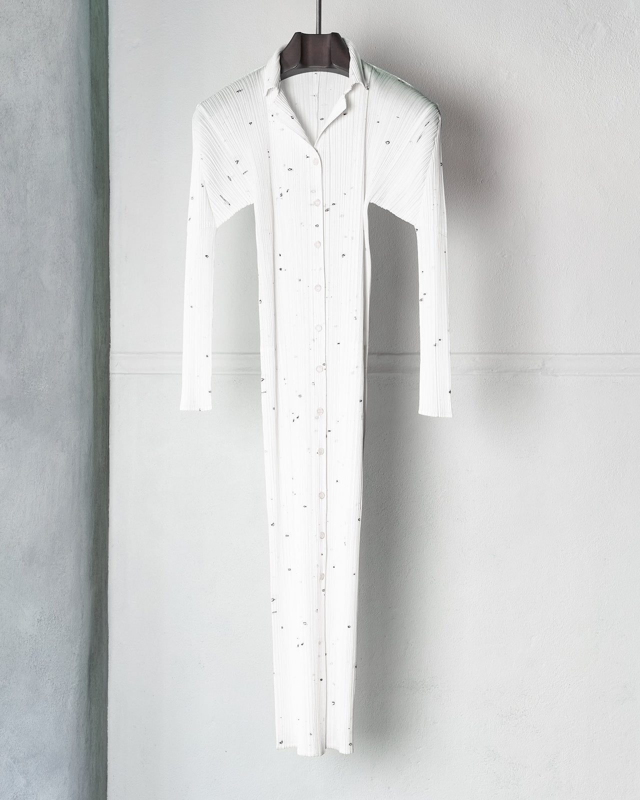 A person in a white, button-up Issey Miyake dress with delicate embellishments stands against a plain background. They wear tall black boots.