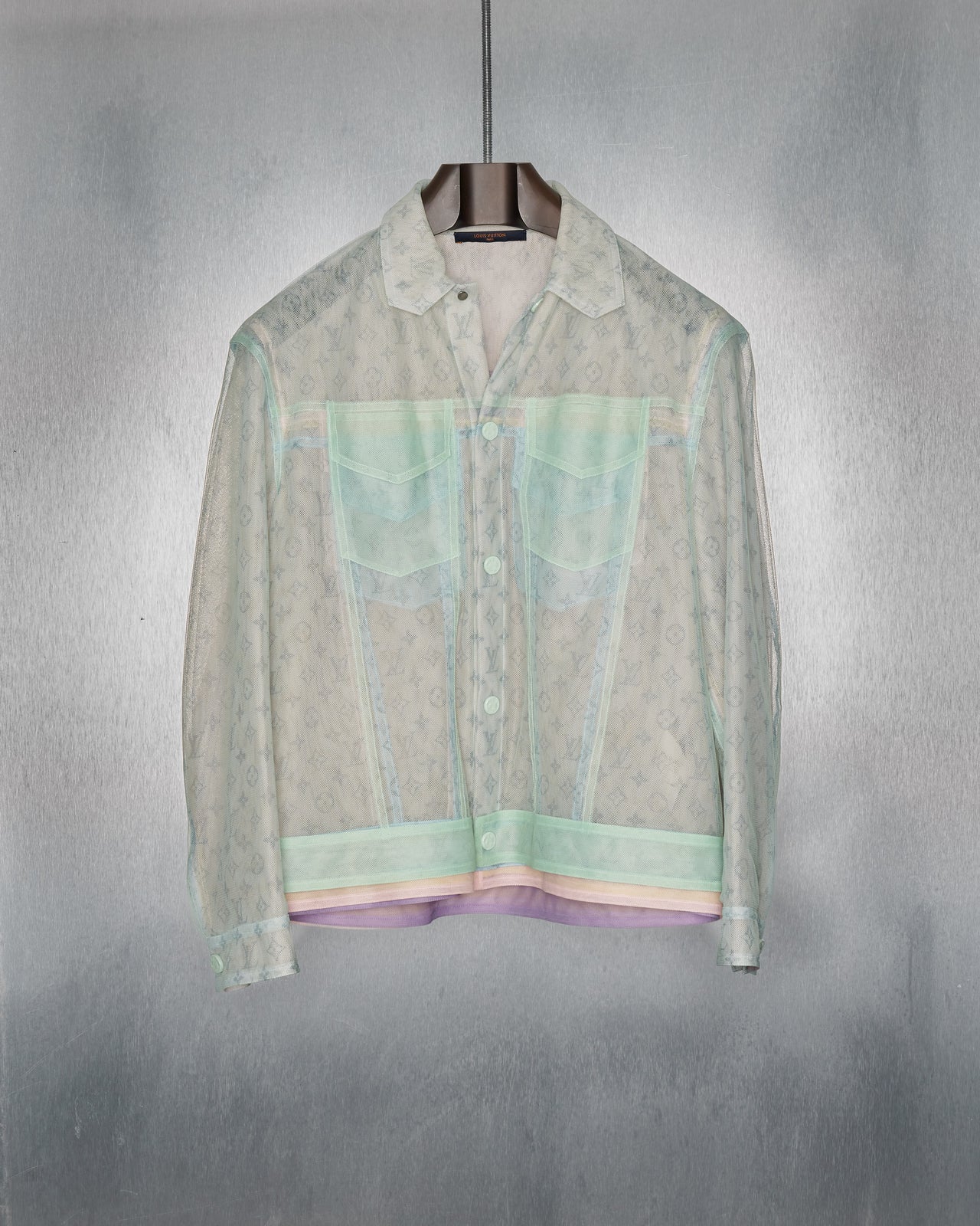 A model wearing a multicoloured, geometric-patterned, layered Louis Vuitton tulle jacket with a trucker fit and a collar, paired with shorts, white sneakers, and accessorised with a necklace.