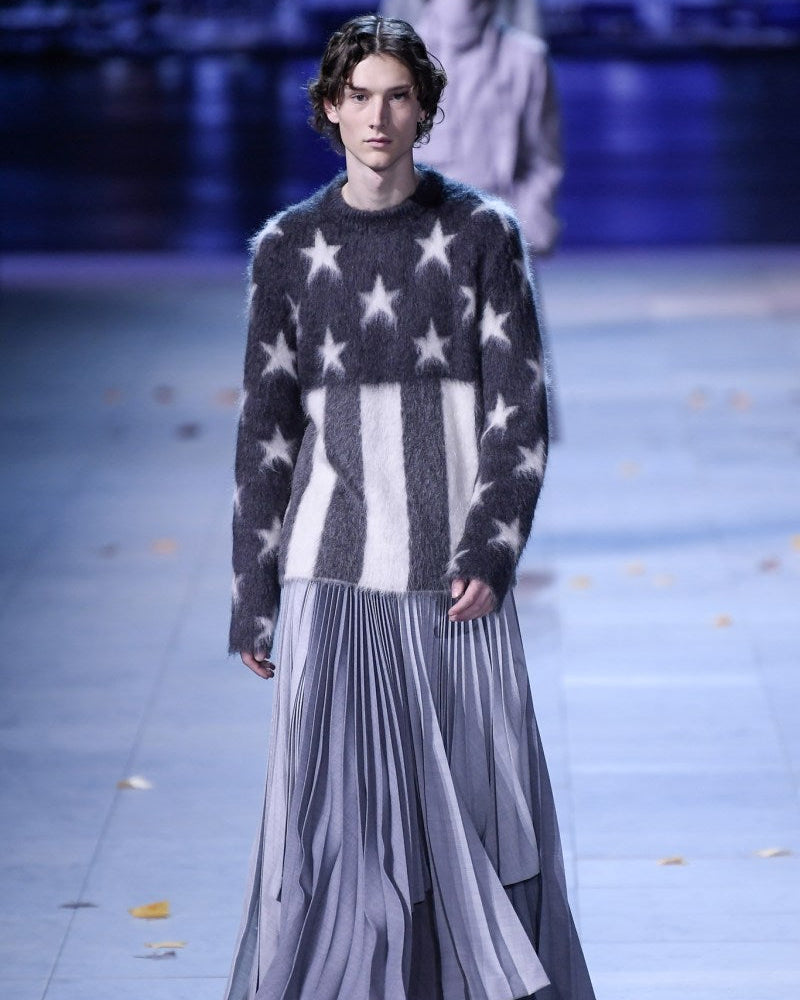 Model walking Louis Vuitton runway in a relaxed, confident manner, wearing a fuzzy sweater with stars and stripes and a flowing grey pleated skirt. The background is softly lit.