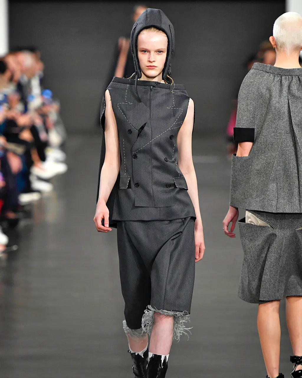 Model walking on a Maison Margiela fashion runway wearing a dark gray outfit with a hood.