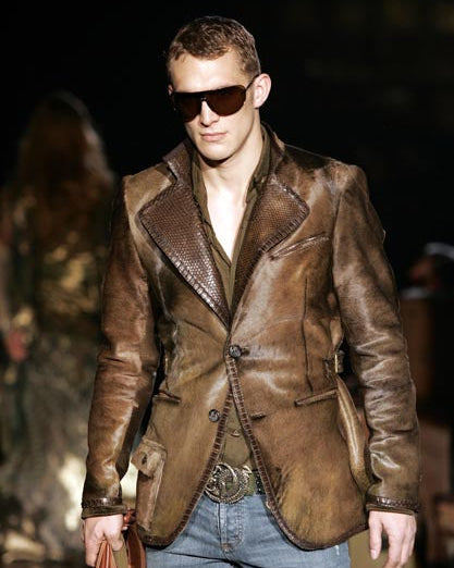 A man in a stylish leather Roberto Cavalli jacket and faded jeans walks confidently down a dimly lit runway, holding a leather bag.