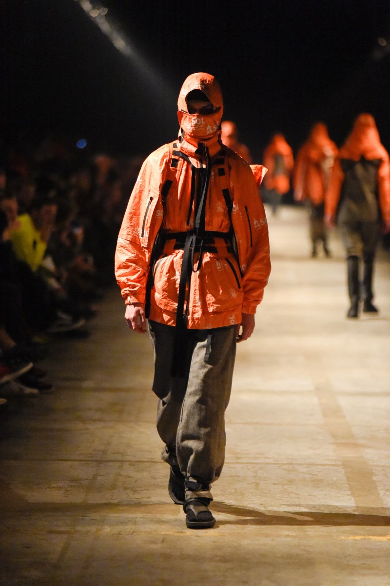 A model wearing an orange hooded jacket by TAKAHIROMIYASHITA TheSoloist and matching face cover walks down a dimly lit runway, exuding a mysterious, futuristic ambiance.