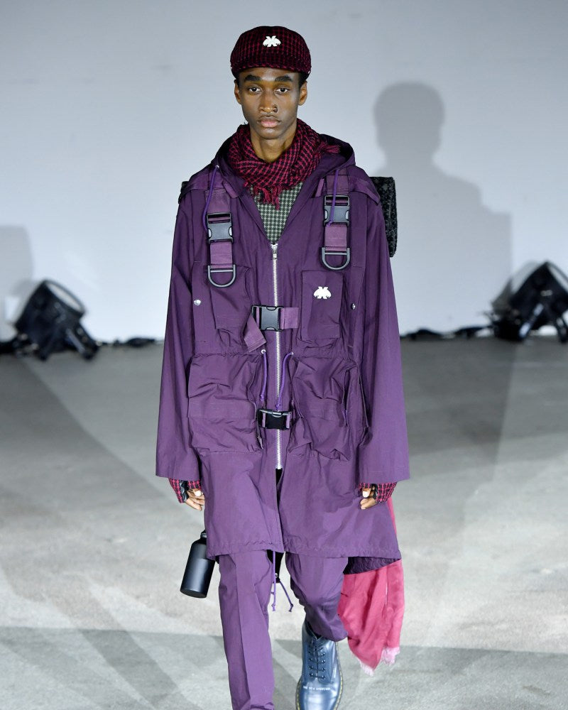 A model walks down the runway in a deep purple, Undercover layered outfit featuring a belted coat and cap, exuding a modern, edgy vibe. Dark boots complete the look.
