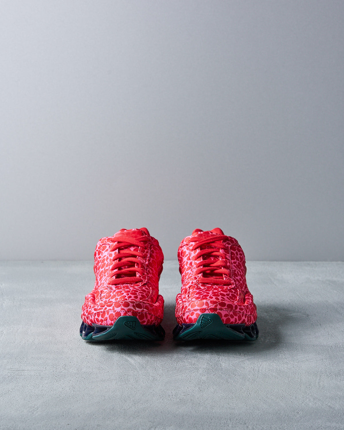 Red sneaker by Adidas x Raf Simons with a floral lace design, featuring unique black coil springs on the sole for a futuristic look. Set against a plain grey background.
