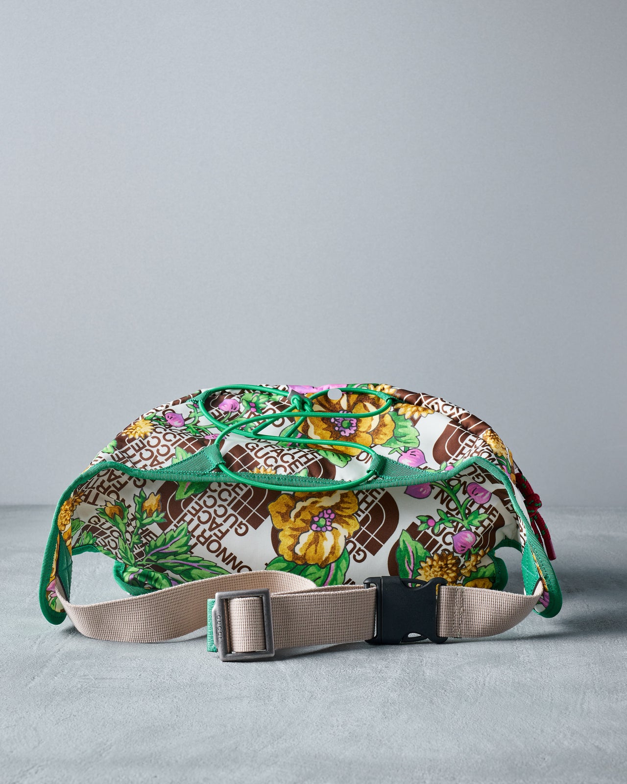 Colourful floral Gucci x The North Face belt bag with bold green trim and tan buckles on a grey surface. Features vibrant purple and yellow flowers with a patterned background.