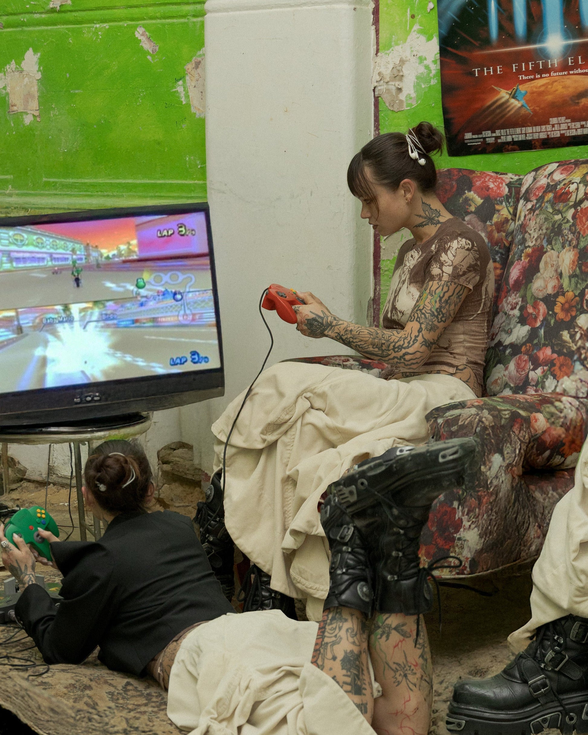 Two people sitting in a room with green walls, a TV, and various items on the floor.