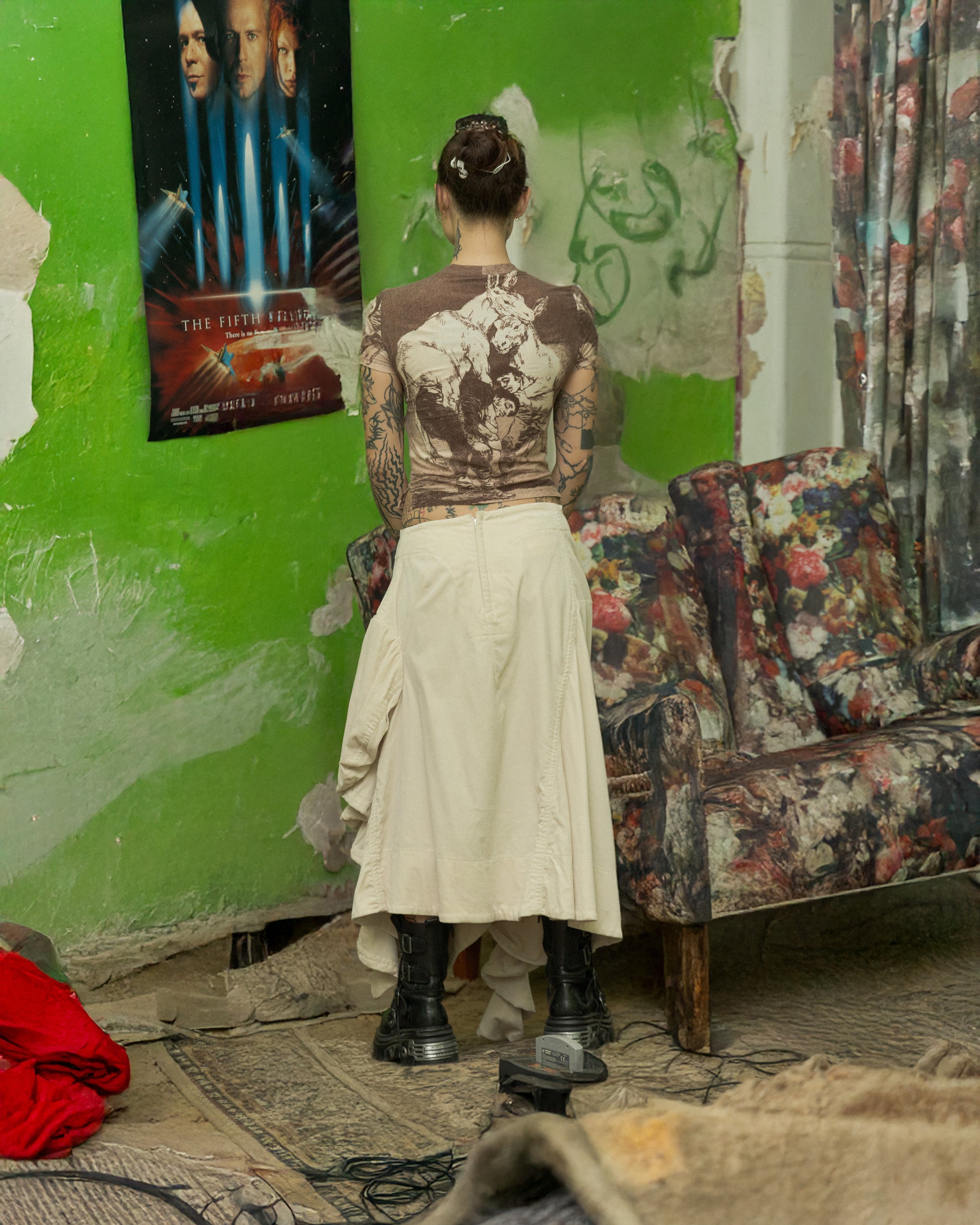 Person with tattoos wears a brown and white Alexander McQueen t-shirt standing in a room with a green wall, floral-patterned sofa, and movie poster.