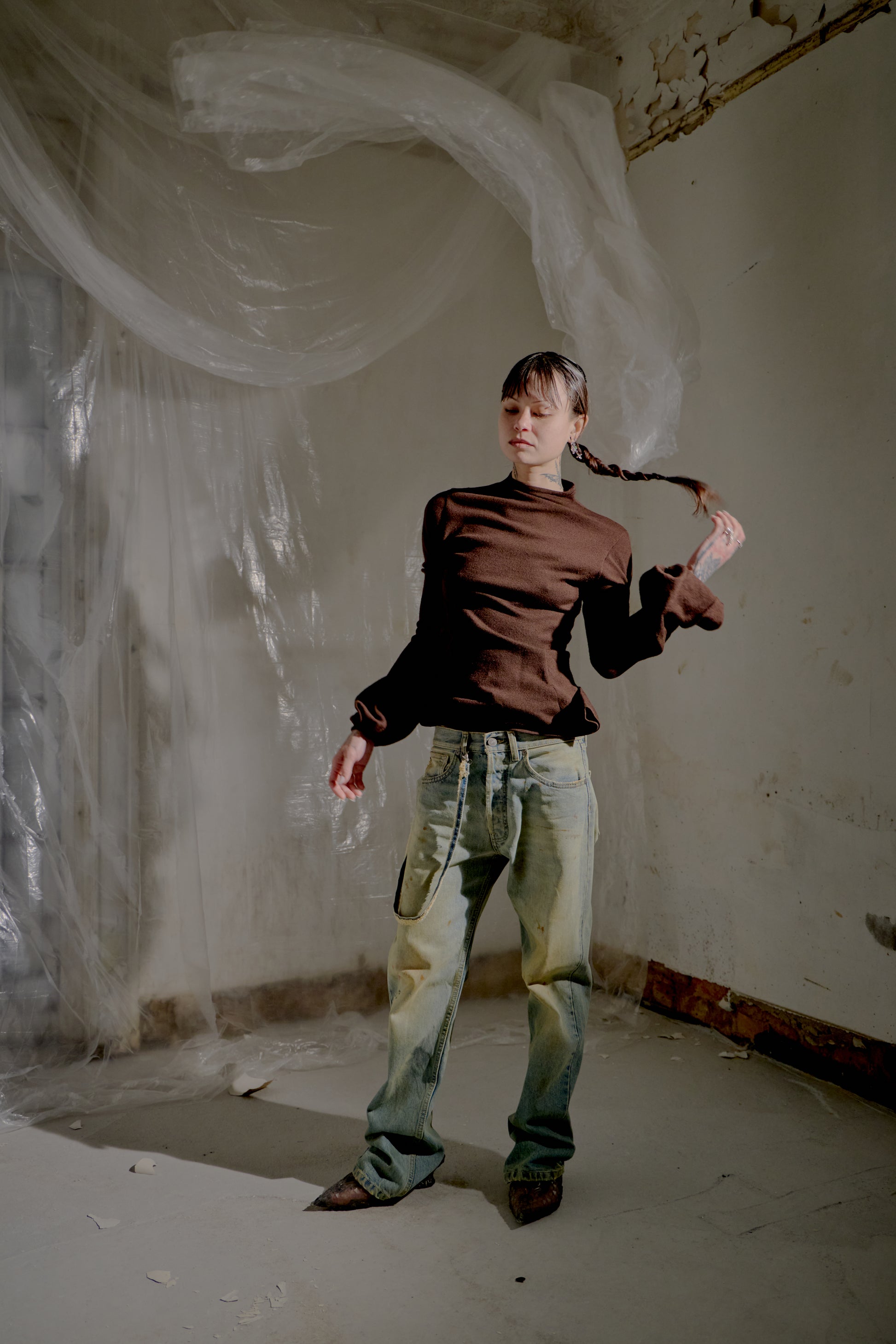 A person with braided hair and eyes closed stands in a dimly lit room, wearing a brown Alexander McQueen top and loose jeans.