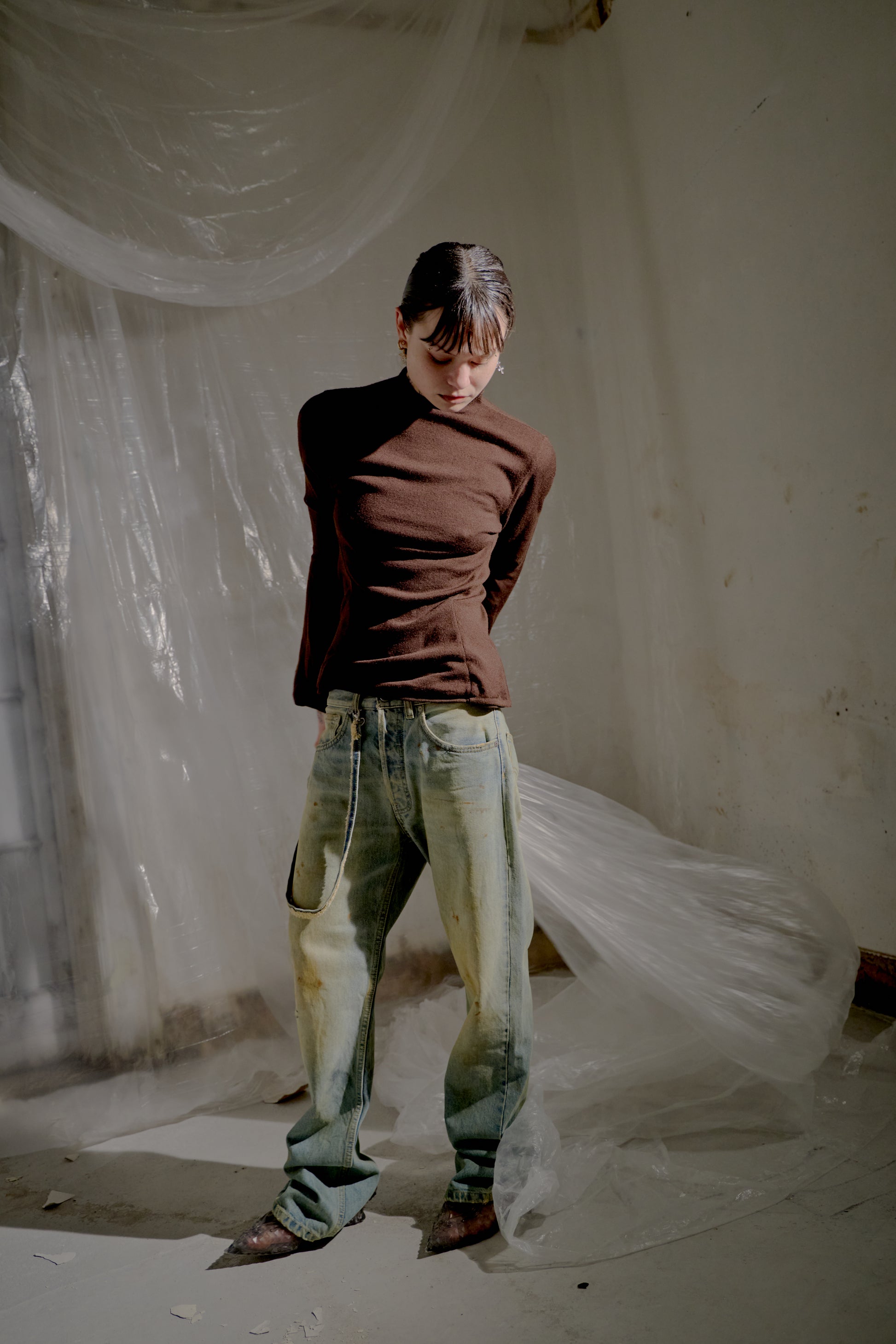 Person wearing a brown Alexander McQueen top and light-coloured jeans standing in a room with draped fabric.