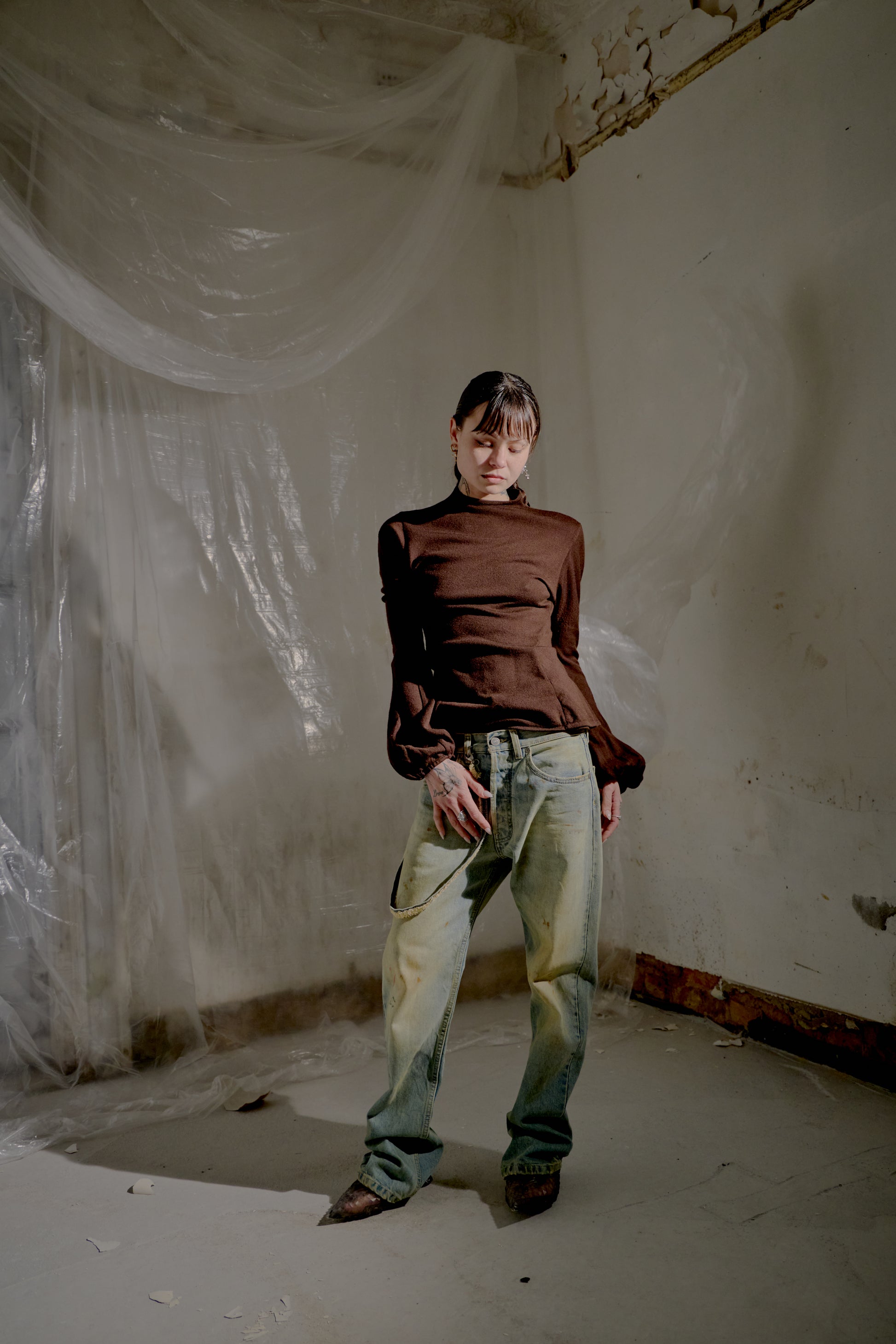 Person standing in a room with white walls and a plastic sheet, wearing a brown  Alexander McQueen top and light blue jeans.