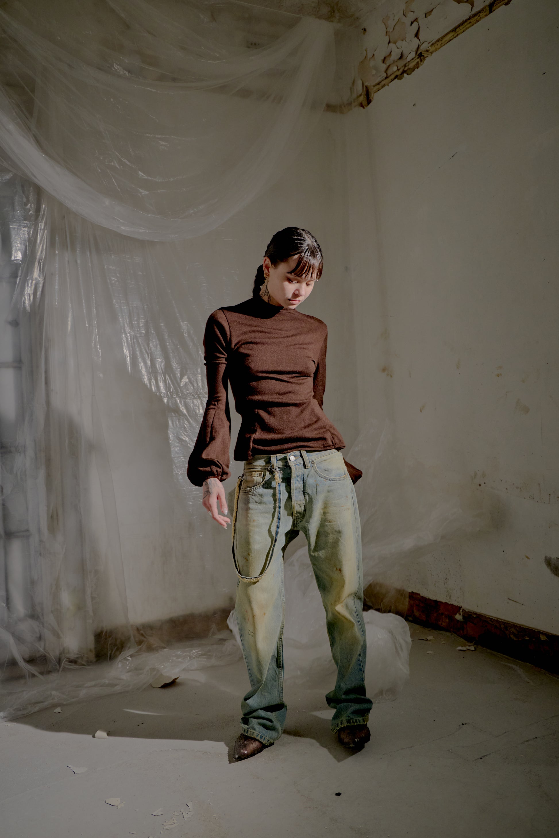 Person wearing a brown Alexander McQueen top and light blue jeans in an abandoned room with white curtains.